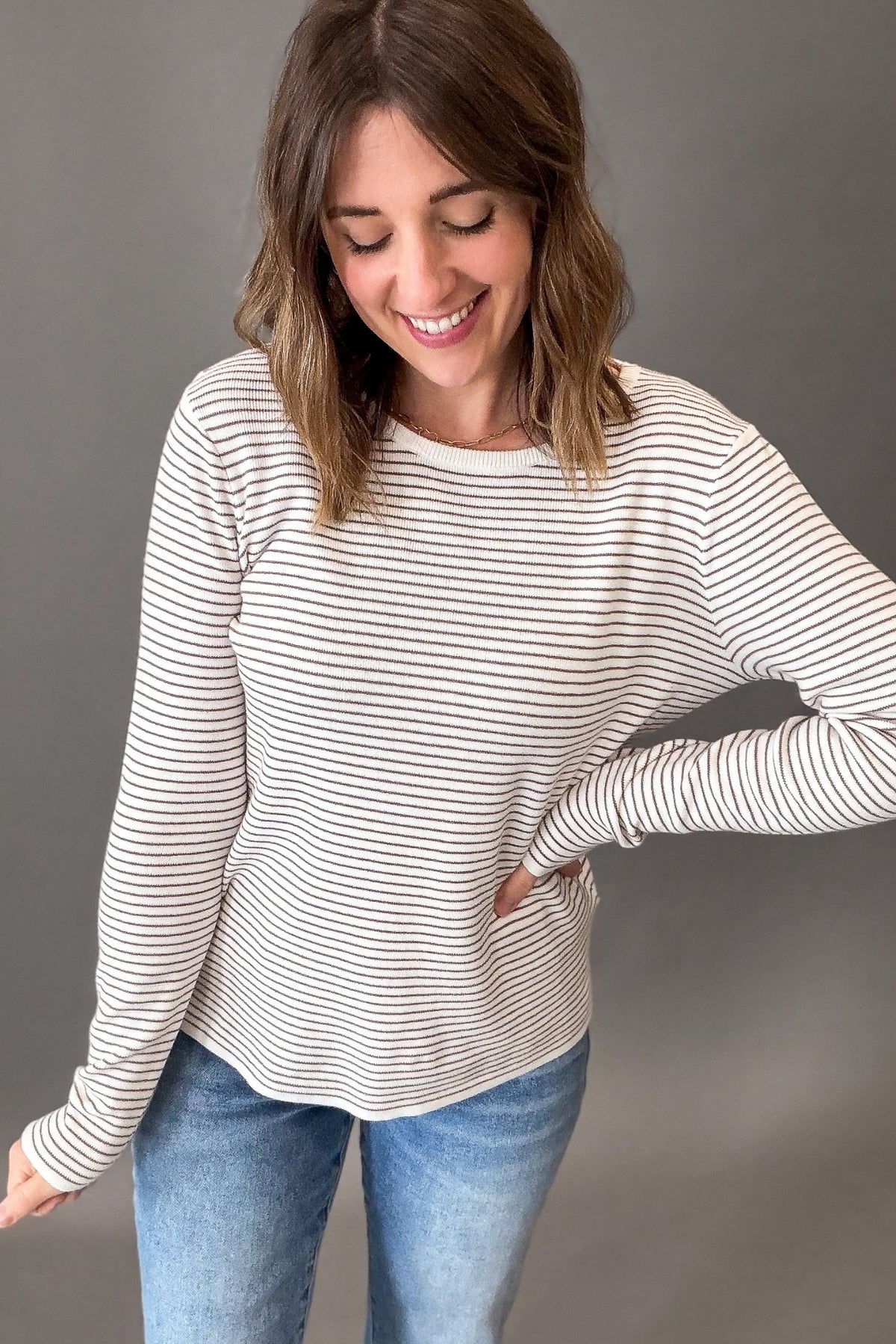 A woman wearing a ivory + mocha horizontally striped long-sleeve top with blue jeans, posing against a solid gray studio background.