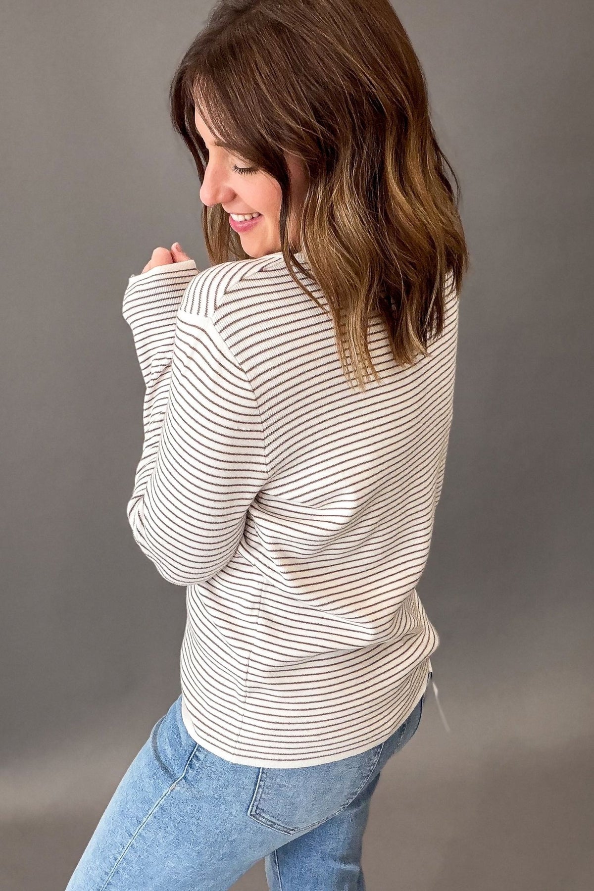 A woman wearing a ivory + mocha horizontally striped long-sleeve top and light blue jeans, shown from a side-back angle against a solid gray studio background.