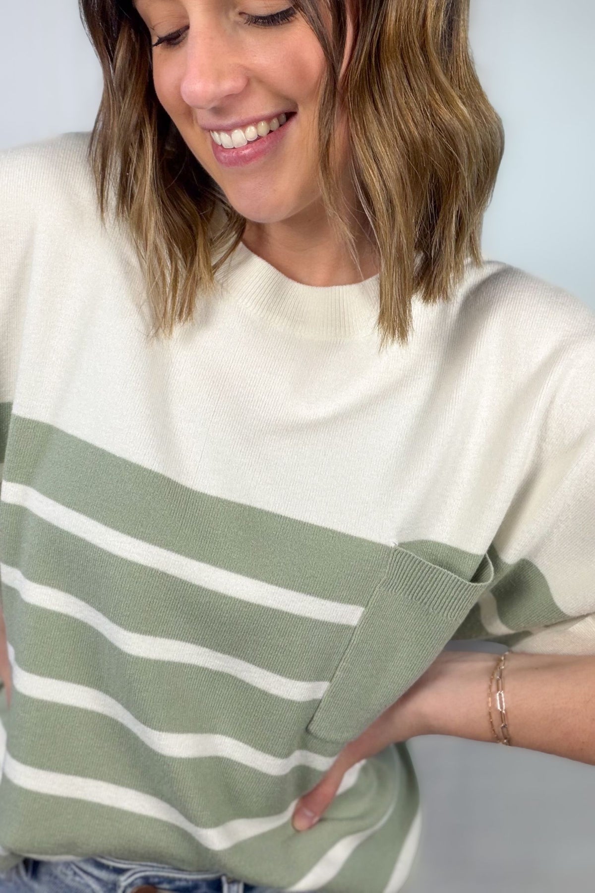 Woman wearing an ivory short sleeve knit top with sage stripes, rolled sleeve cuffs, chest pocket, and light wash jeans showing close up details