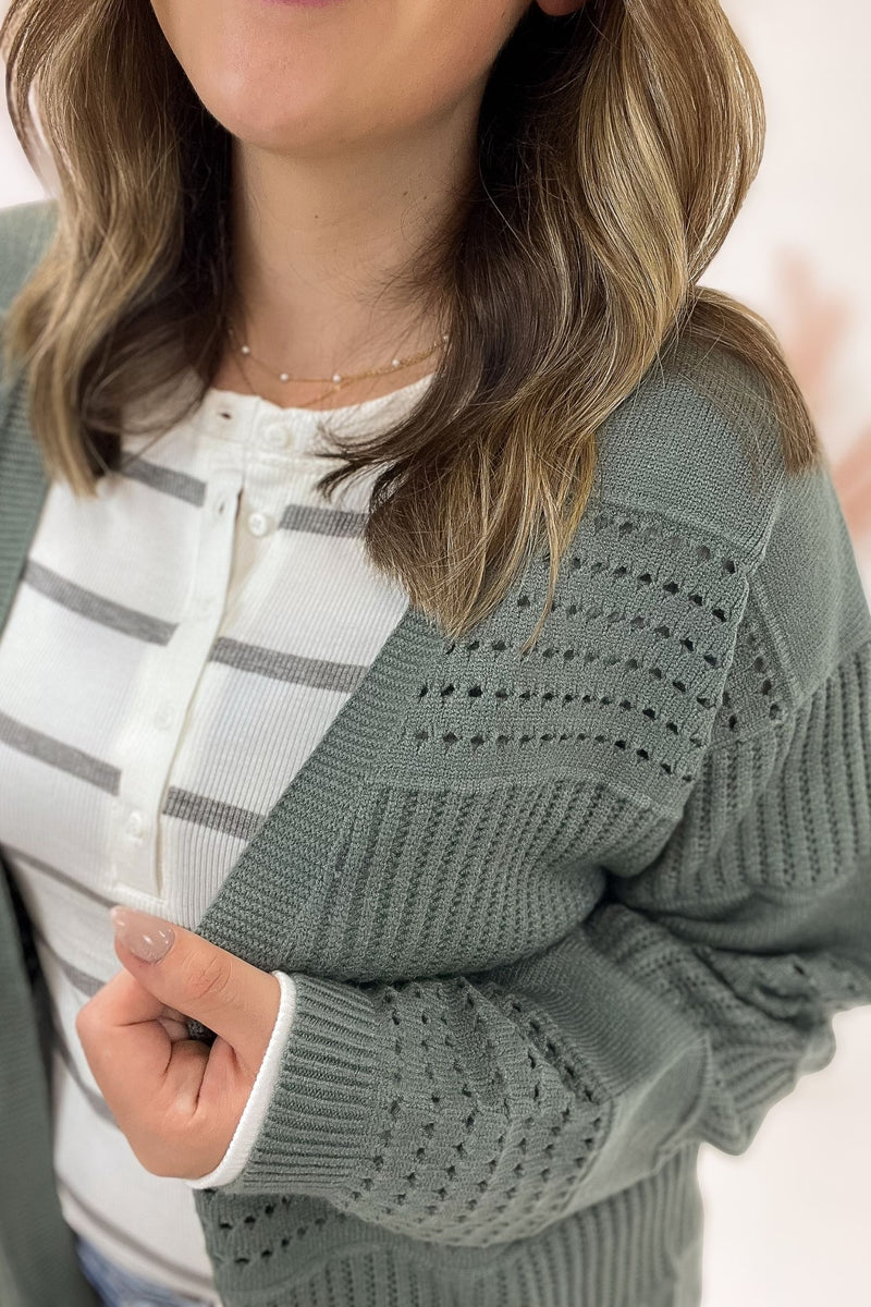 Person wearing a green sweater with a white and gray striped shirt underneath, on a light pink background.
