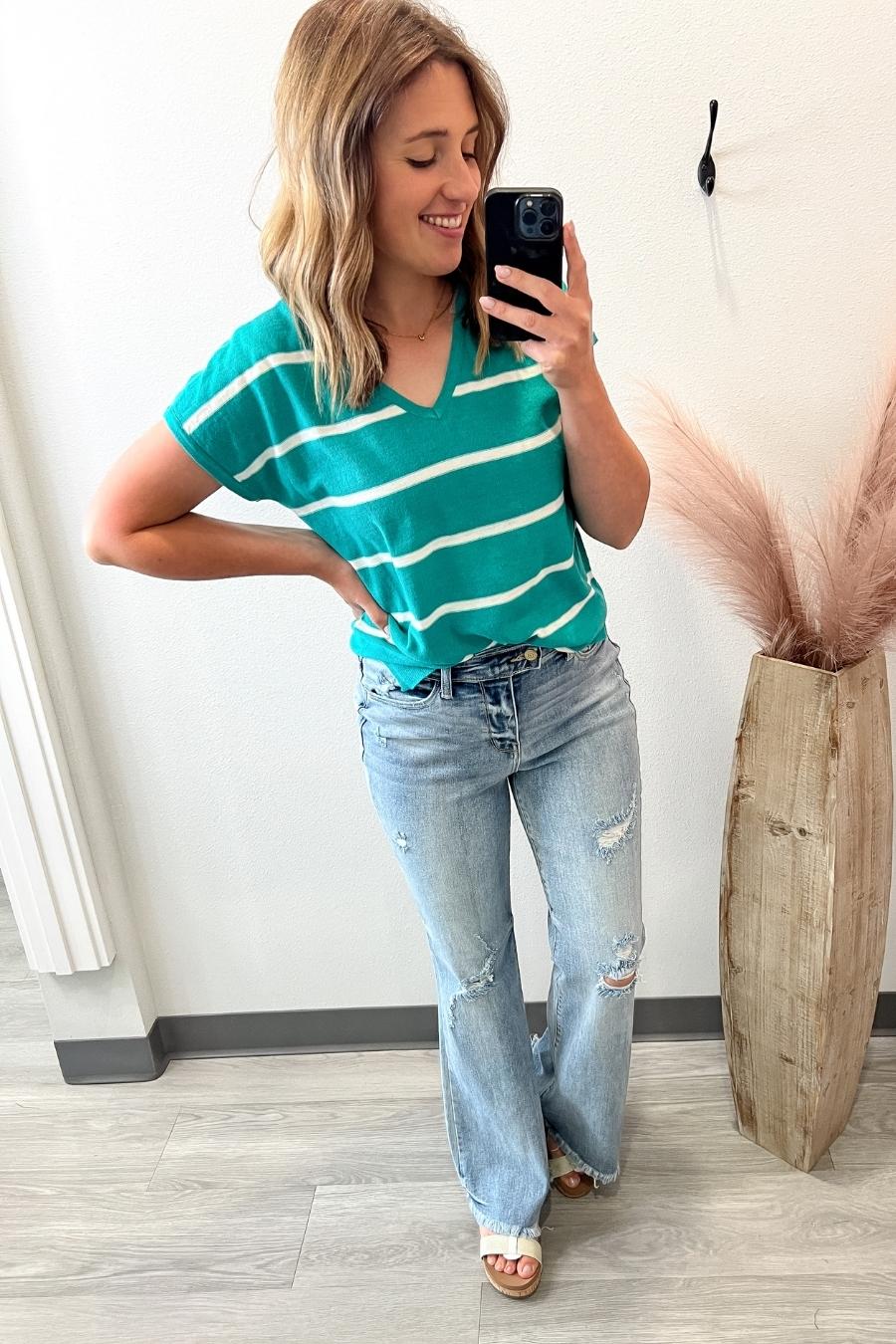 Woman taking a mirror selfie wearing a green striped shirt and light blue jeans, standing next to a decorative plant.