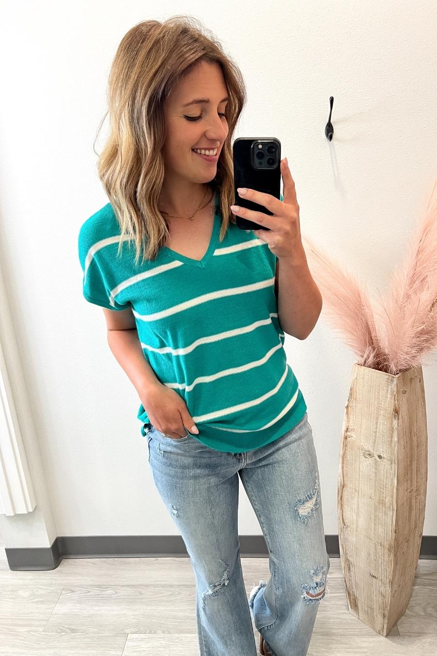 Woman taking a mirror selfie wearing a teal and white striped shirt and light blue jeans.