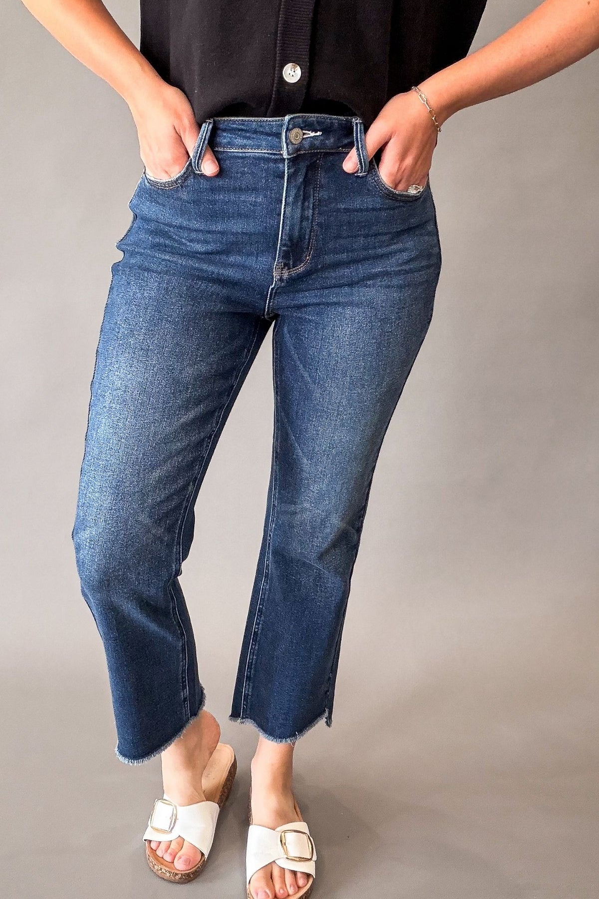 A person wearing a dark wash blue cropped jeans with a raw hem, paired with a black top and white slide sandals, standing against a neutral gray studio background.