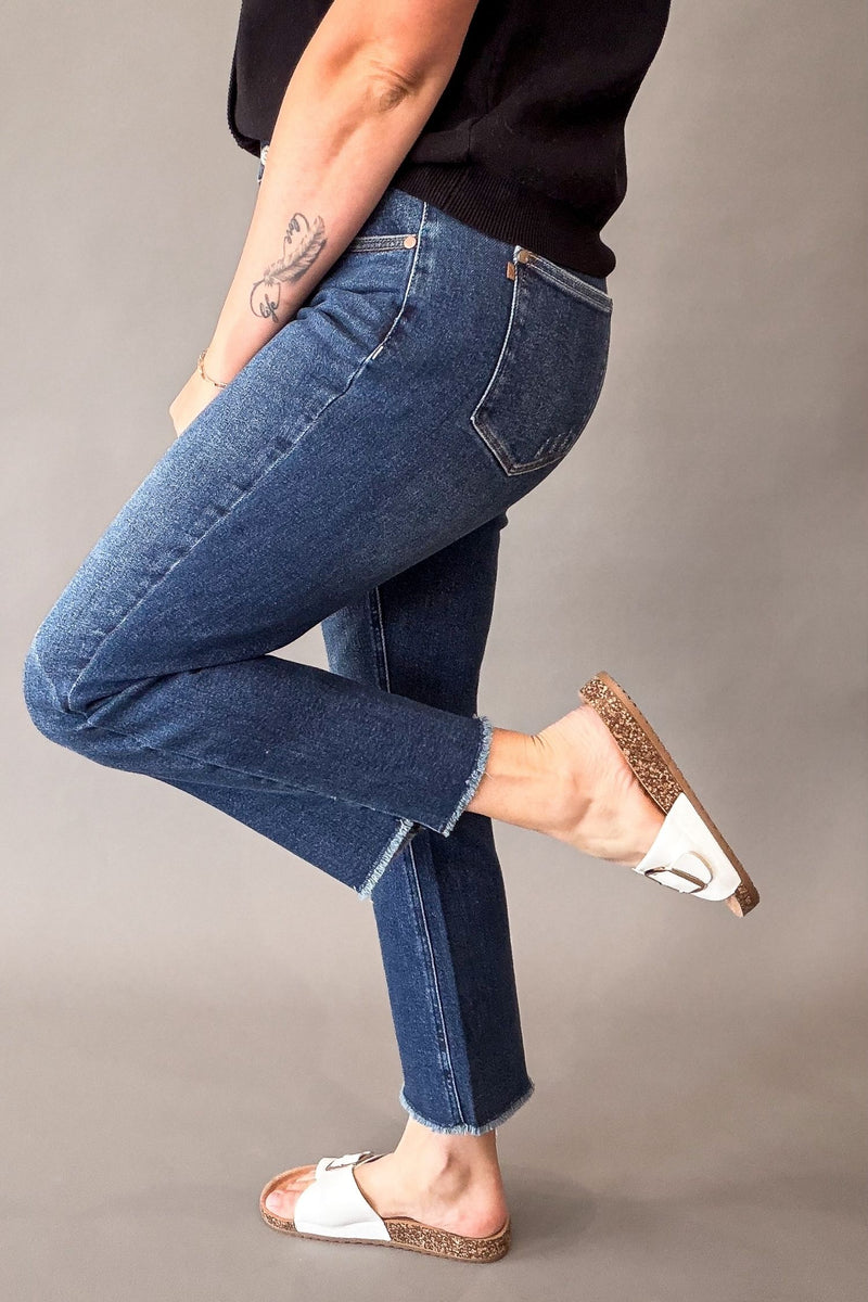 A person wearing blue cropped jeans with a raw hem, paired with a black top and white slide sandals, shown from the waist down against a plain gray studio background.