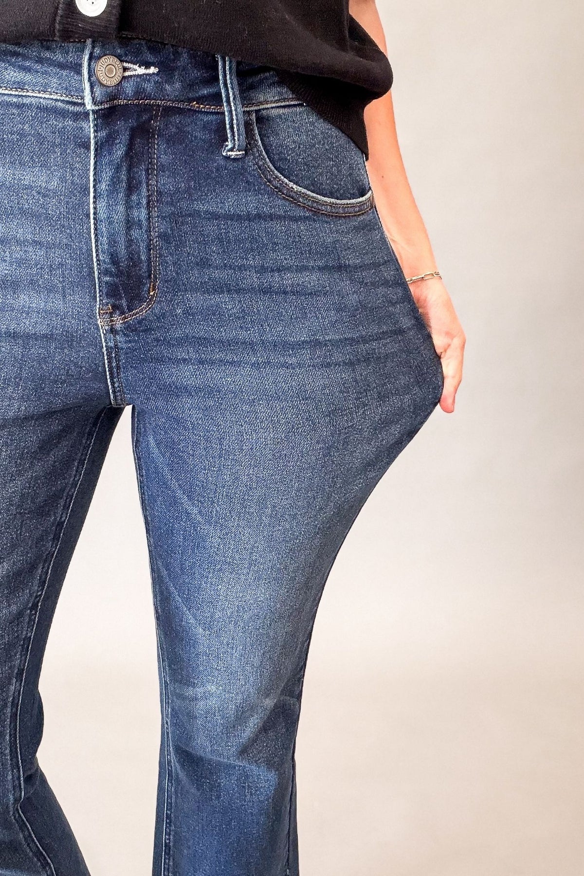 Close-up of a person wearing dark-wash blue denim jeans, showing the front waistband, button, belt loops, pocket, and the side stretch of the denim fabric.
