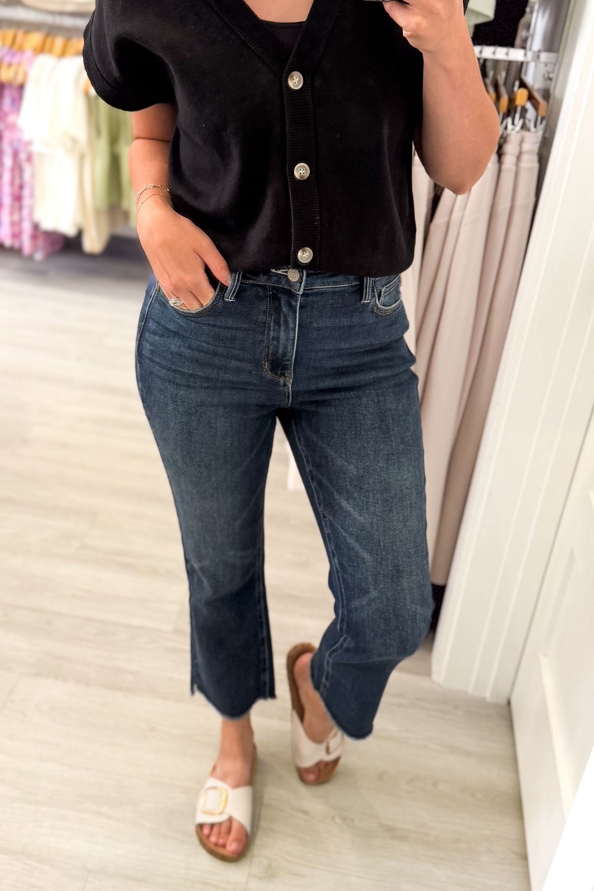 A person wearing dark blue cropped flare jeans, a black short-sleeve button-up top, and white slide sandals, standing in a clothing store.