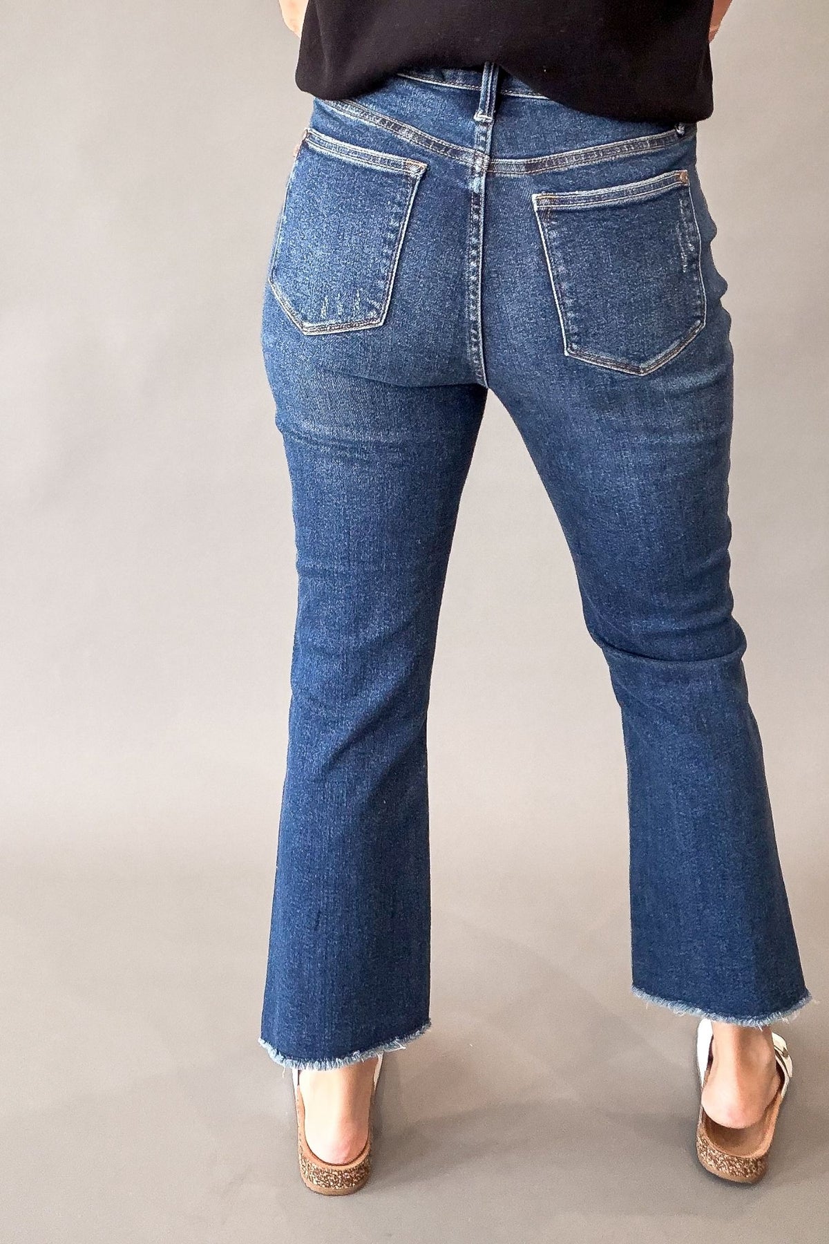 Rear view of a person wearing blue denim kick flare jeans with a frayed hem and white sandals, against a plain light gray studio background.