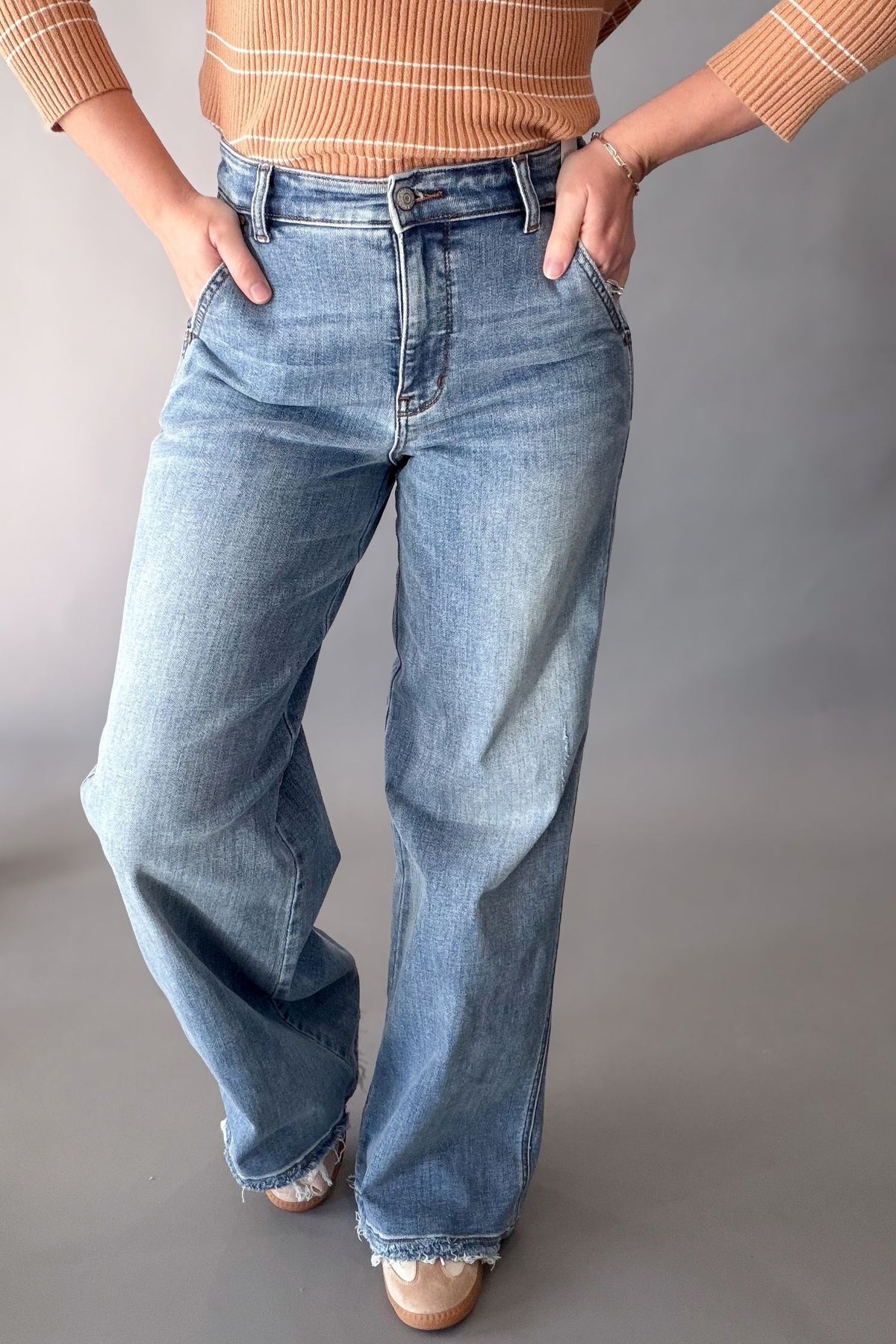 A person wearing light-wash blue wide-leg jeans, paired with a tan striped knit top and beige shoes, standing against a plain gray studio background.