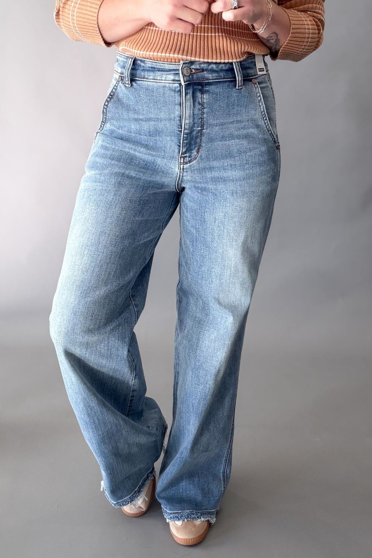 A person wearing light blue wide-leg denim jeans with a faded wash, paired with a rust-orange ribbed sweater and beige pointed shoes, standing against a neutral gray studio background.