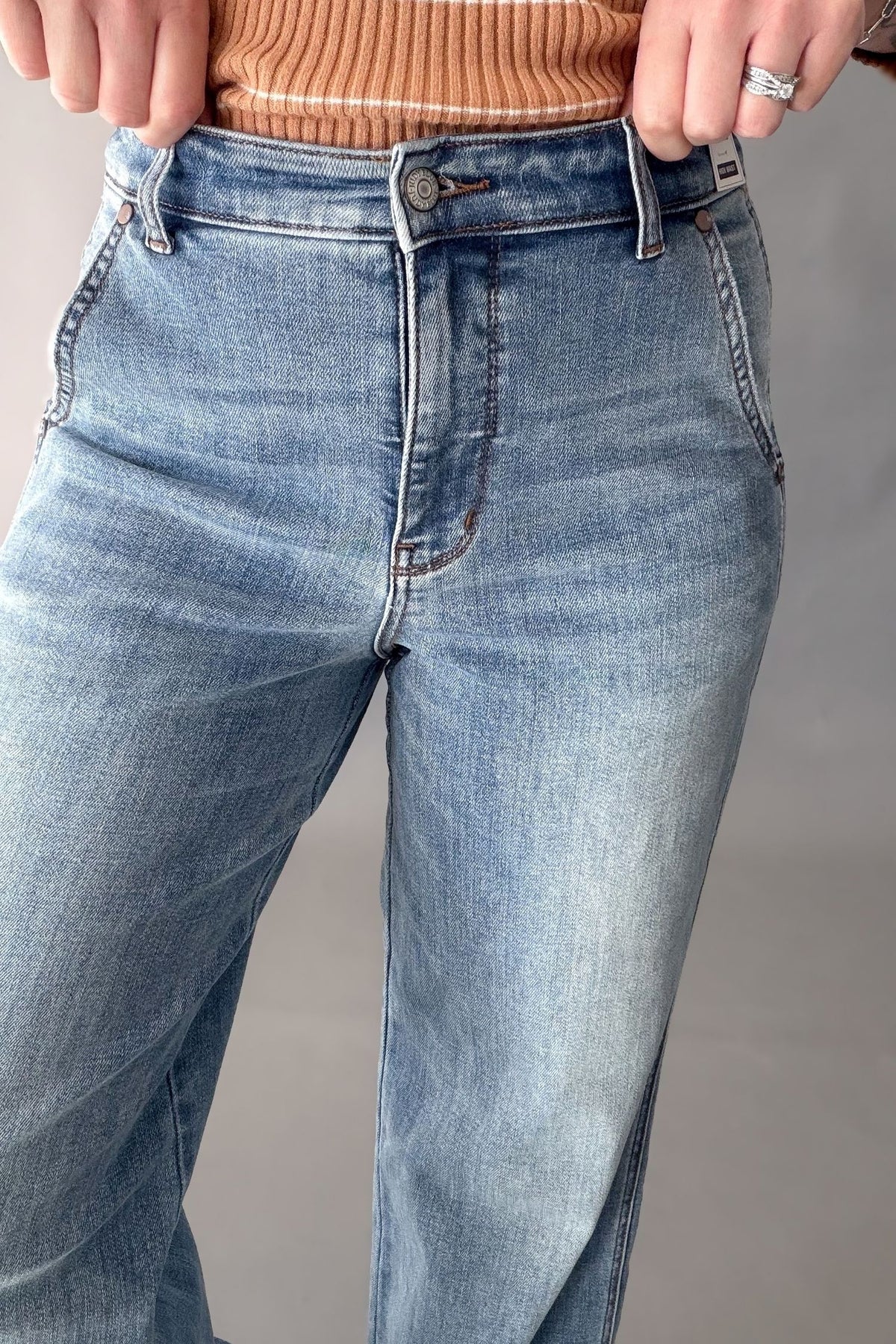 Close-up of a person wearing light blue high-rise denim jeans, showing the front waistband, button, belt loops, and upper legs against a plain gray background.