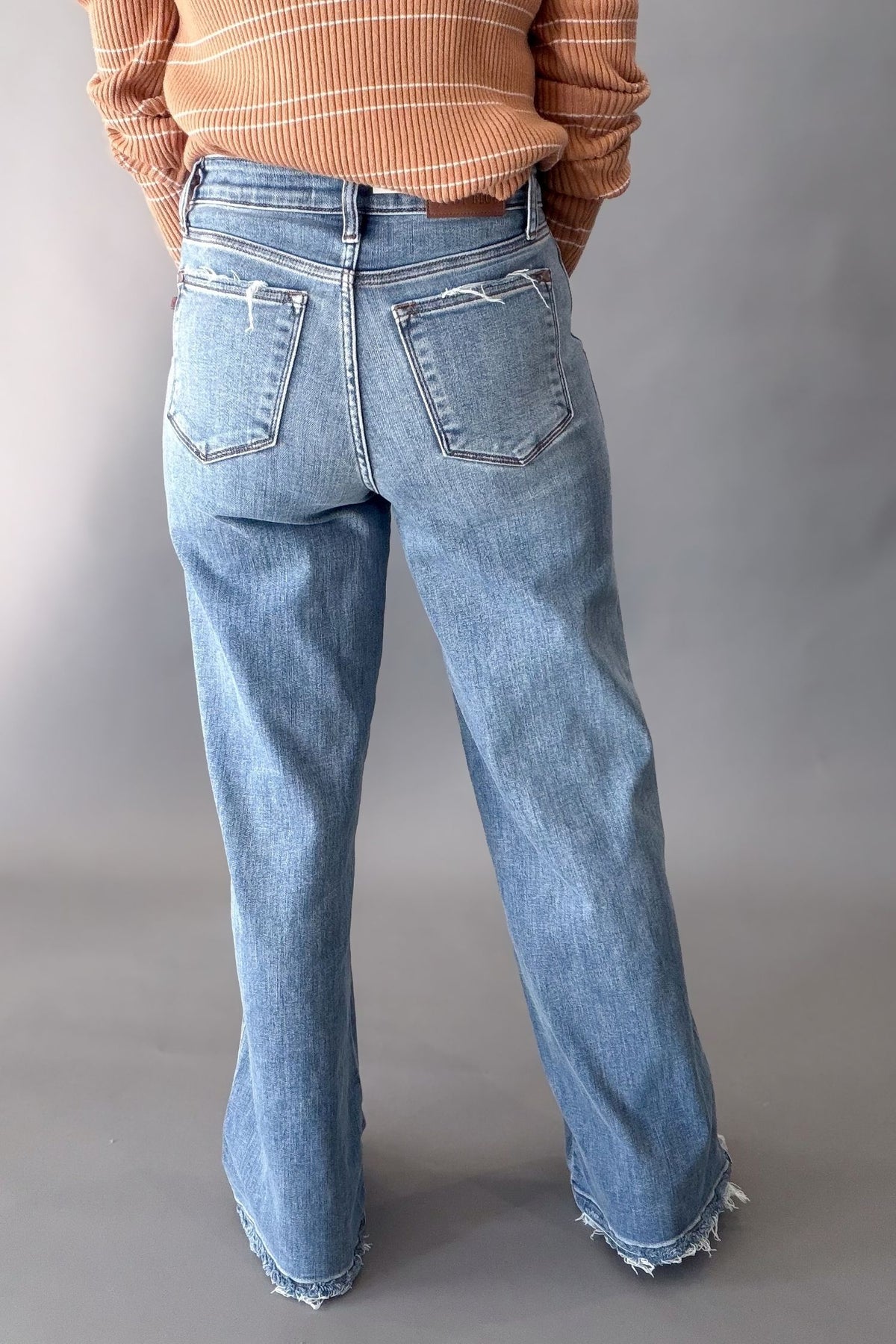 A person shown from the back wearing light-wash blue wide-leg jeans with frayed hems and distressed details on the back pockets, paired with an orange striped long-sleeve top against a gray studio background.