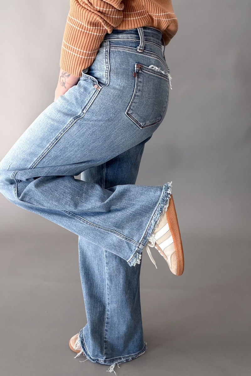 A person wearing light-wash blue flare jeans with frayed hems, paired with a brown striped long-sleeve top and brown-and-white sneakers, photographed against a plain gray studio background.