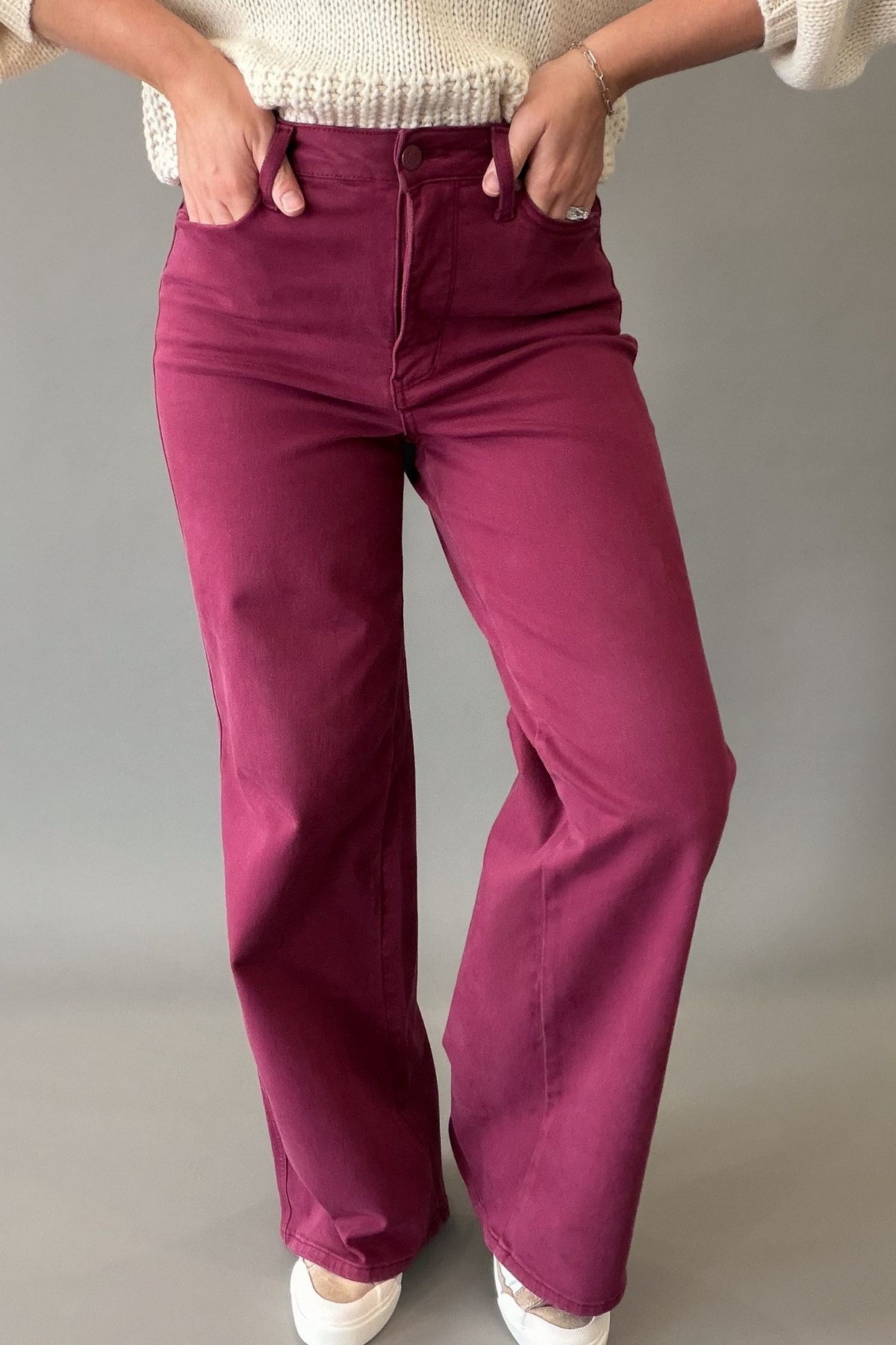 A person wearing high-waisted, wide-leg pants in a boysenberry color, paired with a cream knit sweater and white sneakers, standing against a neutral gray studio background.