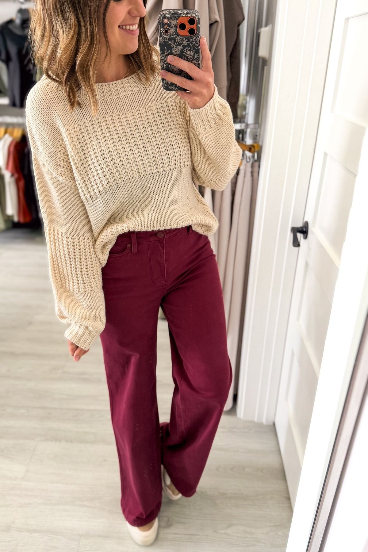 A woman takes a mirror selfie in a fitting room or boutique, wearing a cream-colored chunky knit sweater and boysenberry wide-leg pants.