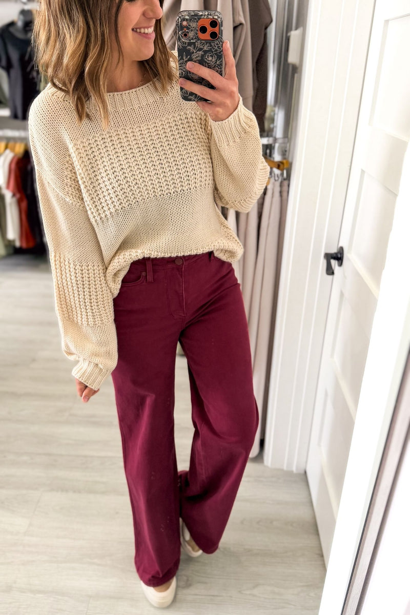 A woman takes a mirror selfie in a fitting room or boutique, wearing a cream-colored chunky knit sweater and boysenberry wide-leg pants.