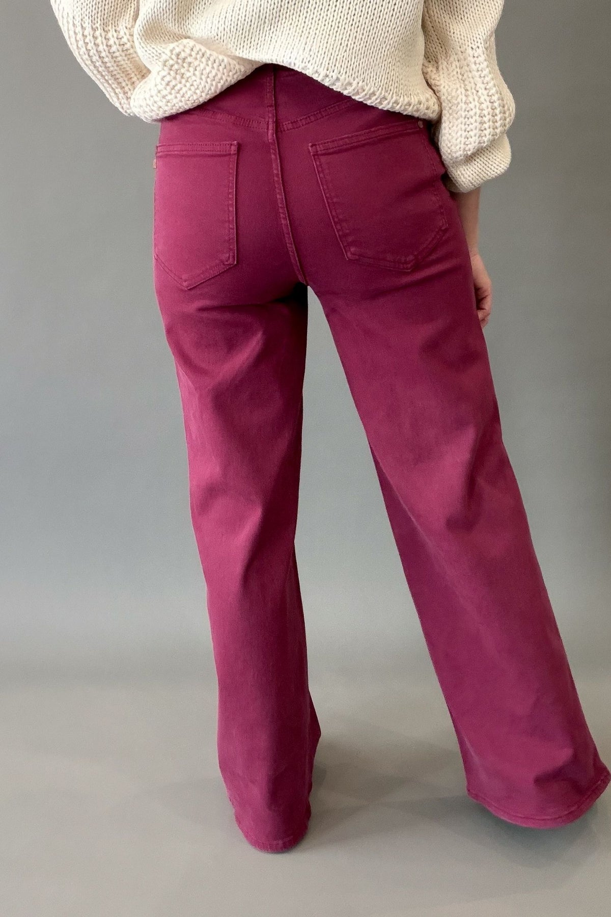A person shown from the back wearing boyesenberry colored wide-leg jeans and a cream knit sweater against a plain gray studio background.