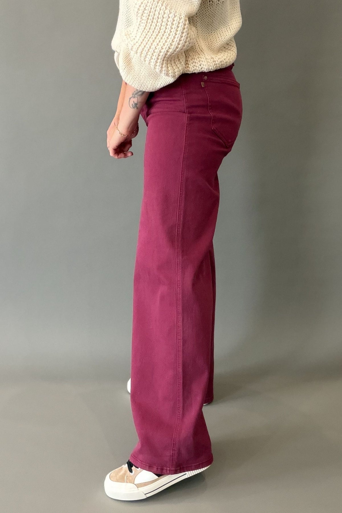 A person shown from the waist down wearing a pair of wide-leg, straight-leg style trousers in a boysenberry color, paired with a cream knit sweater and white sneakers, against a plain gray studio background.
