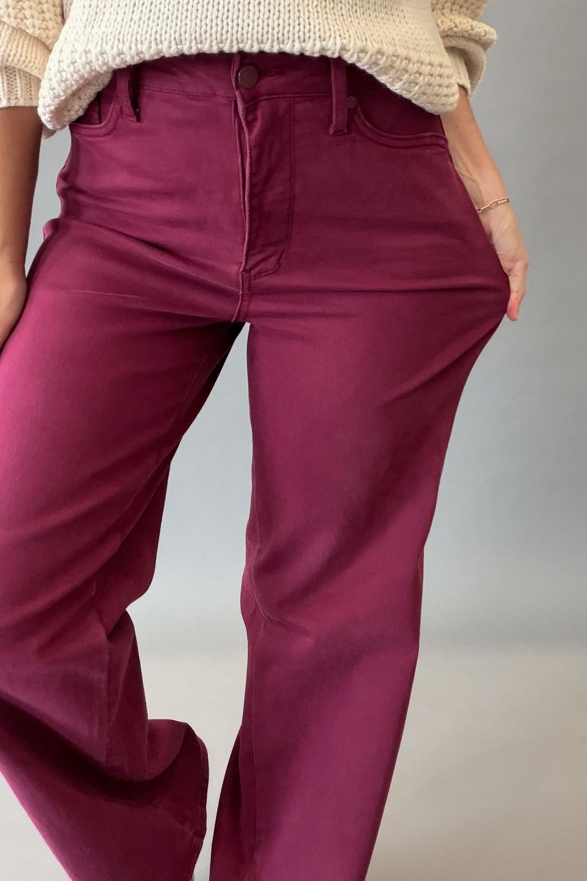 A person wearing high-waisted, wide-leg corduroy pants in a boysenberry color, paired with a cream knit sweater, shown from the waist down against a neutral gray background.
