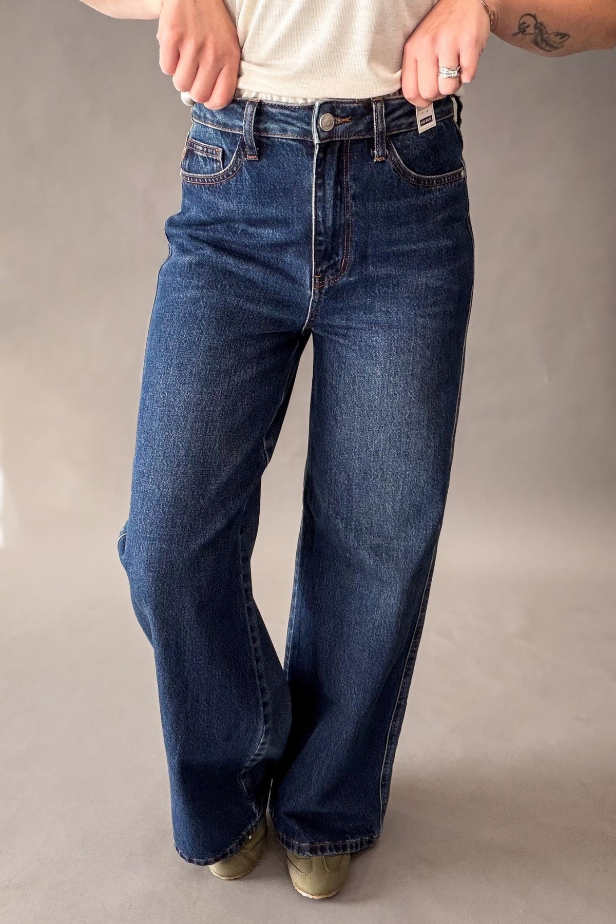 A person wearing dark blue wide-leg denim jeans with a faded wash, paired with a cream t-shirt and olive green sneakers, shown from the waist down against a neutral gray studio background.
