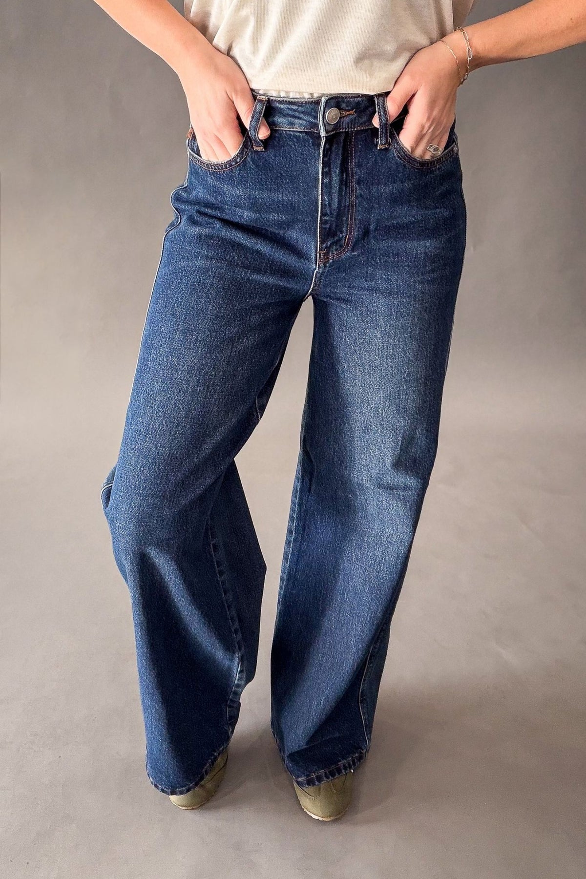 A person wearing a pair of wide-leg blue denim jeans with a faded wash, paired with a light-colored top and sage green sneakers, standing against a neutral gray studio background.