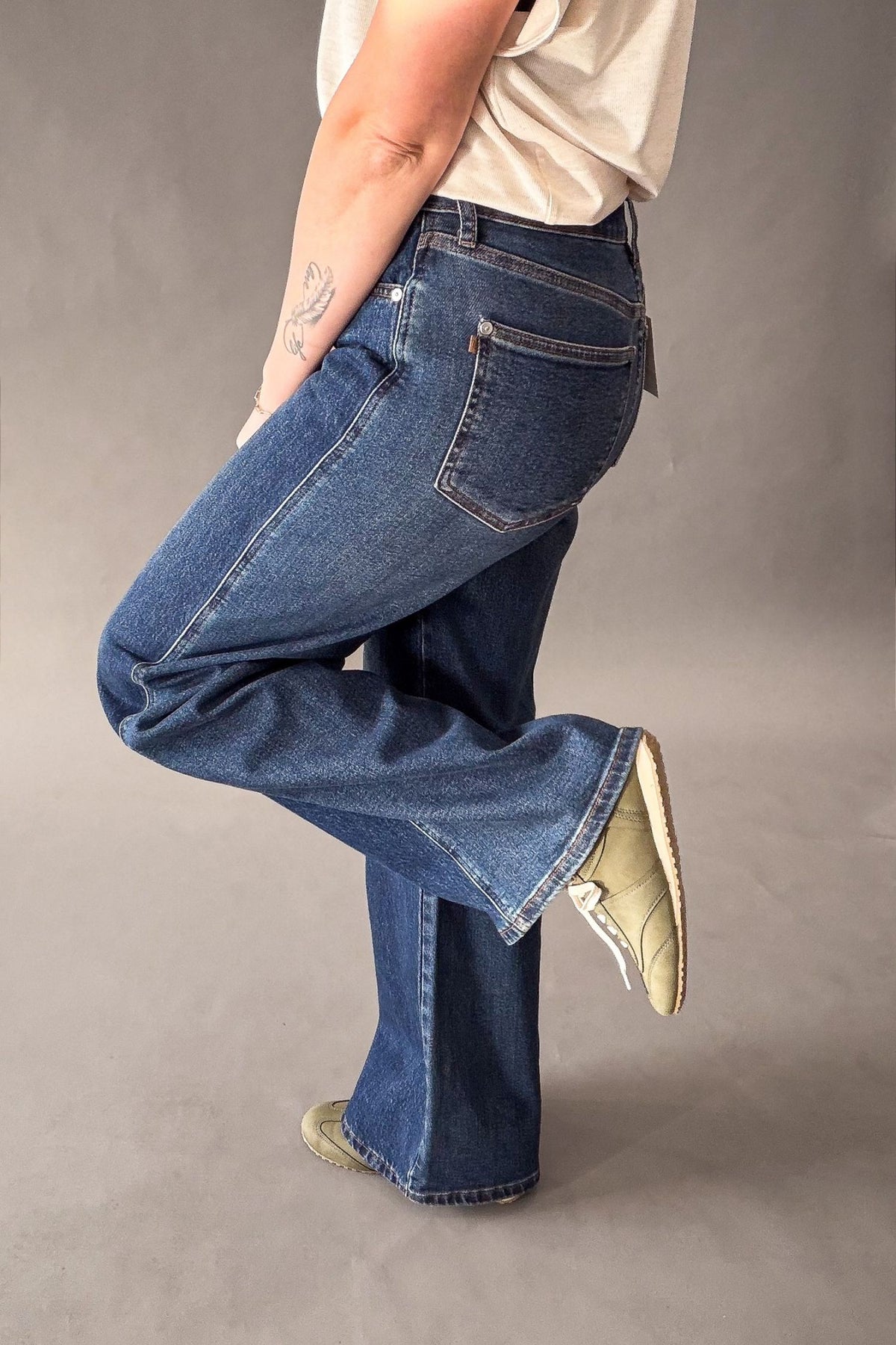 A person wearing dark-wash blue wide leg jeans, a cream t-shirt, and olive sneakers, shown from the waist down in a studio setting with a gray background.
