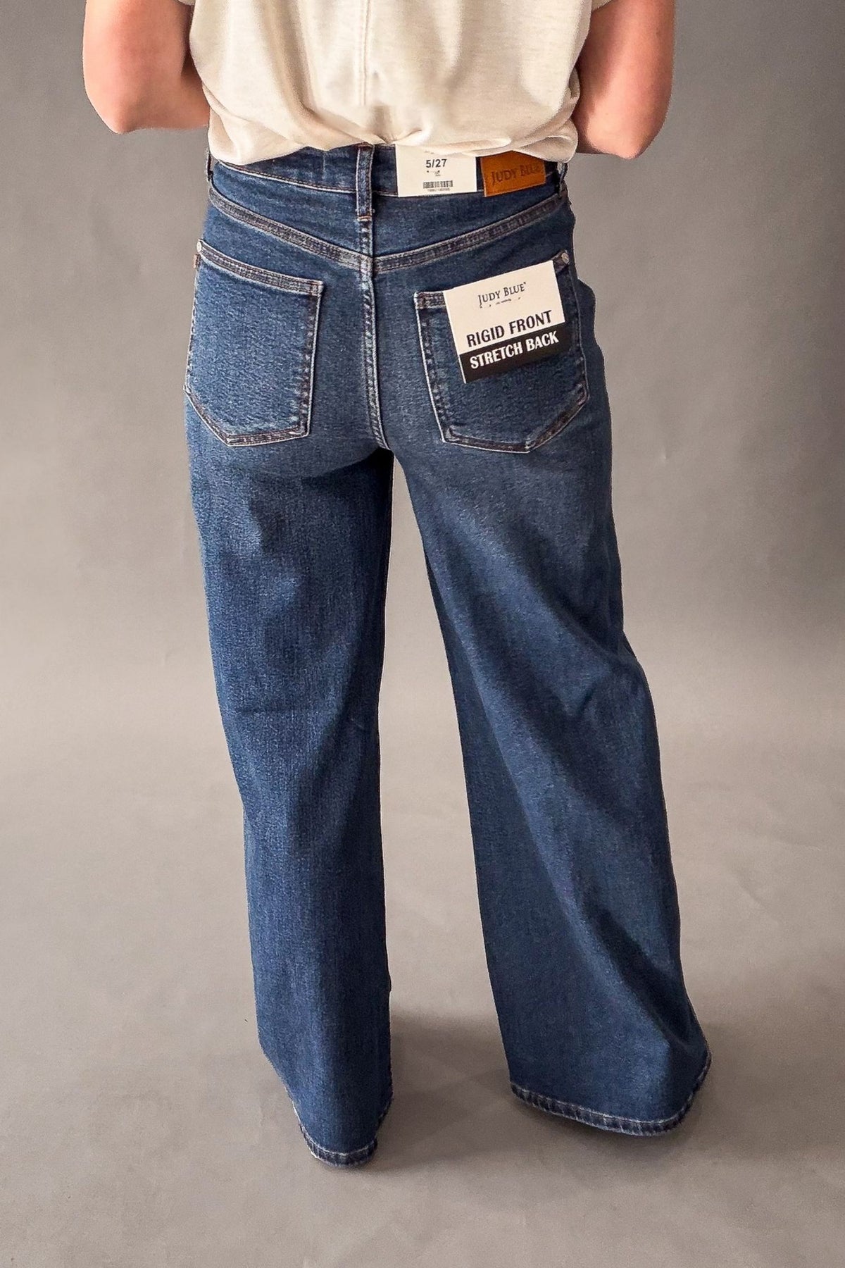 A person shown from the back wearing blue wide-leg jeans with a cream t-shirt, photographed against a neutral gray studio background. The back pocket of the jeans features a white patch reading 'Rigid front, STRETCH BACK'.