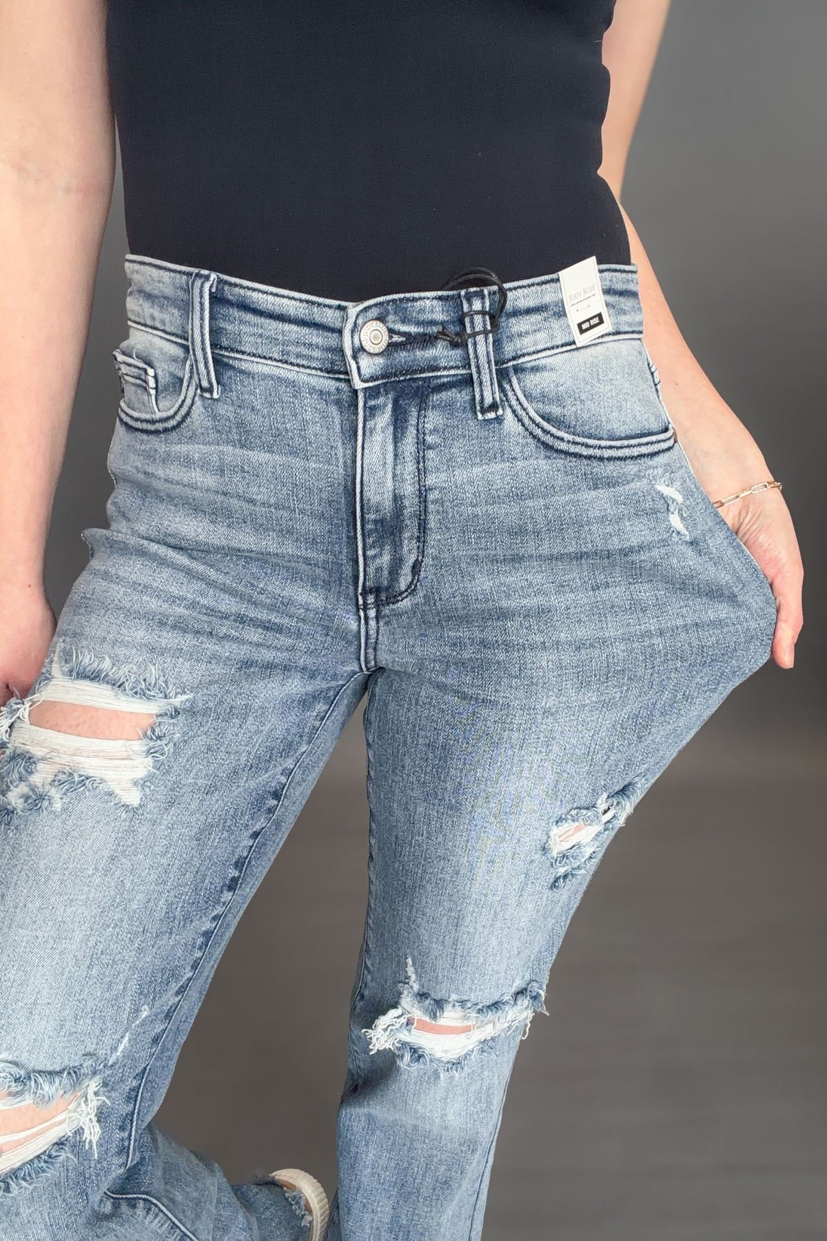 Woman wearing Judy Blue Sandra Destroyed Mid Rise Straight with a black tank and white sneakers on a grey background showing a closeup of the jean details