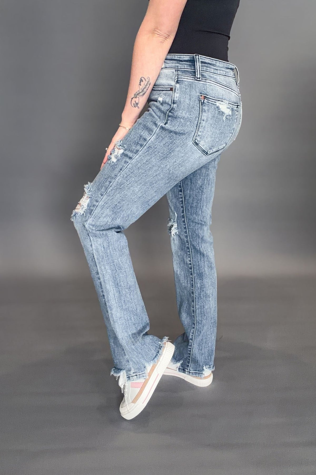 Woman wearing Judy Blue Sandra Destroyed Mid Rise Straight with a black tank and white sneakers on a grey background showing the side view of the denim jeans