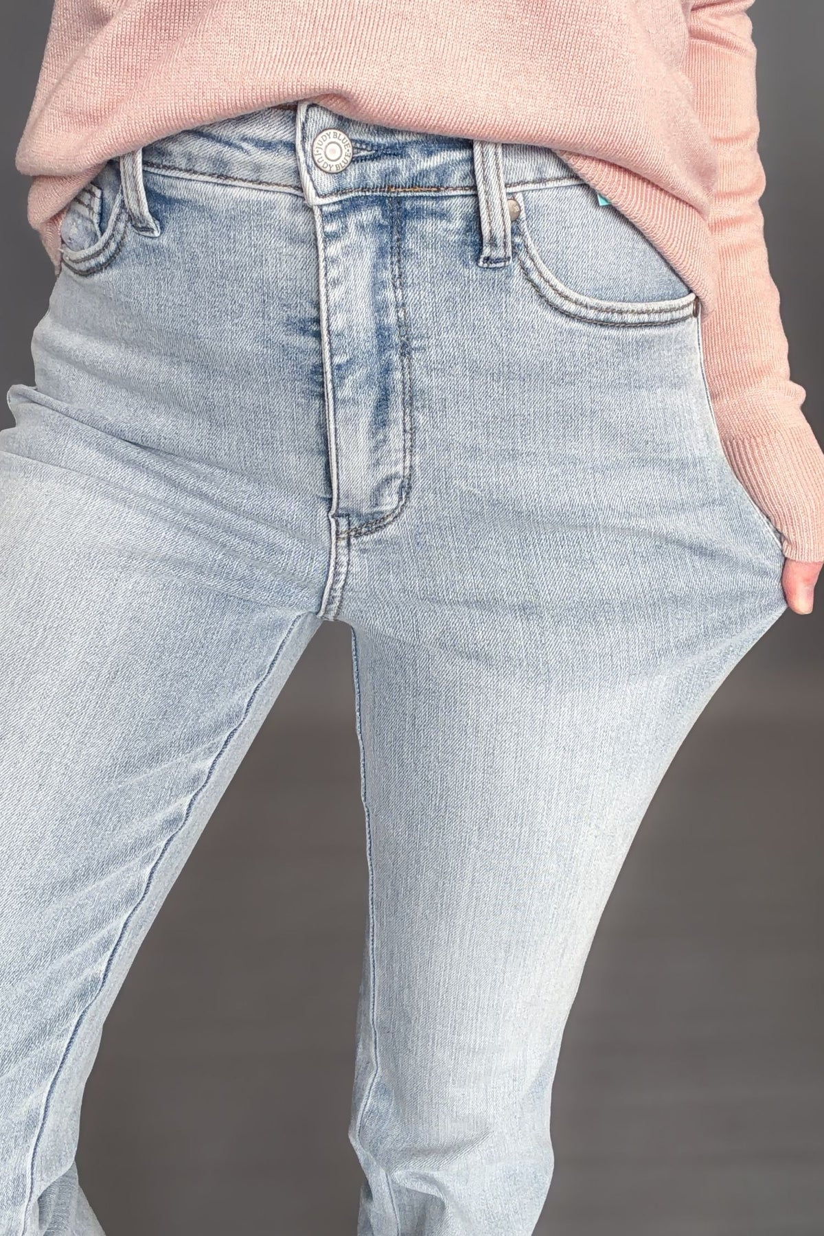 Woman wearing Judy Blue Tessa High Waist Tummy Control Bootcut with a light pink long sleeve with white sneakers on a dark grey background showing a detailed view and stretch
