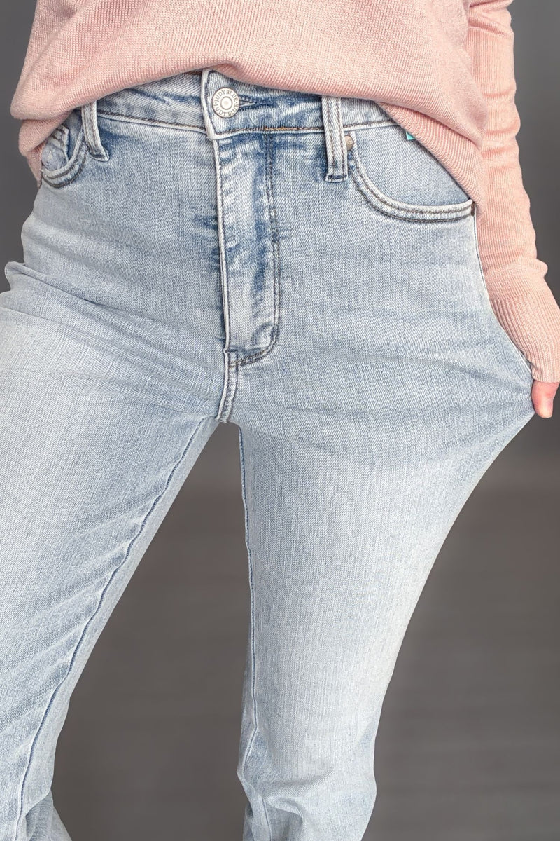 Woman wearing Judy Blue Tessa High Waist Tummy Control Bootcut with a light pink long sleeve with white sneakers on a dark grey background showing a detailed view and stretch