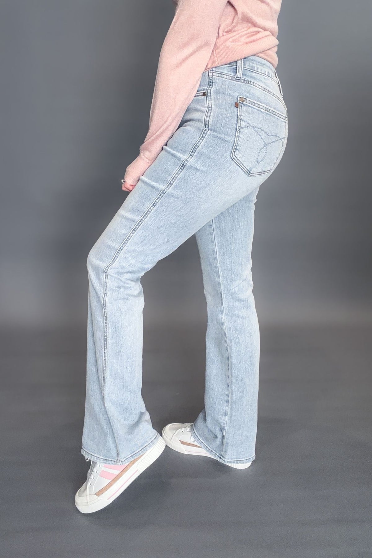 Woman wearing Judy Blue Tessa High Waist Tummy Control Bootcut with a light pink long sleeve with white sneakers on a dark grey background showing the side view of the denim