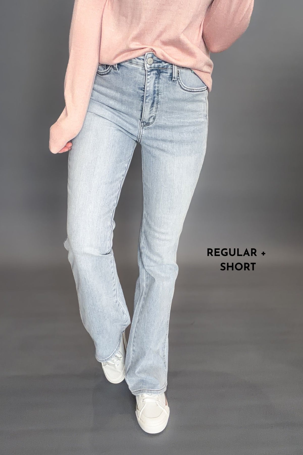 Woman wearing Judy Blue Tessa High Waist Tummy Control Bootcut with a light pink long sleeve with white sneakers on a dark grey background with the words regular and short