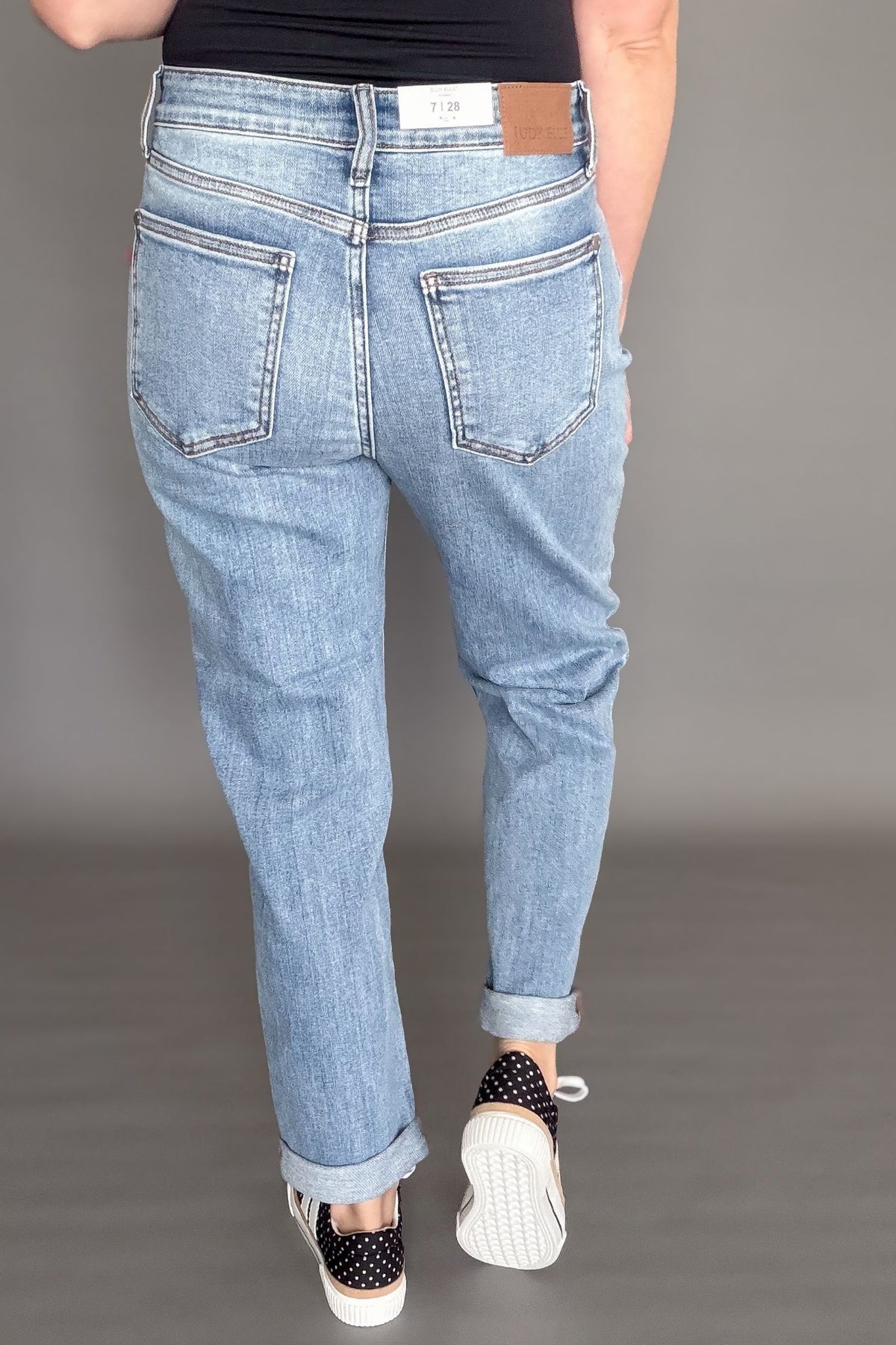 Woman wearing Judy Blue mid rise boyfriend jeans in light wash denim styled cuffed with black sneakers showing the back view of the jeans.
