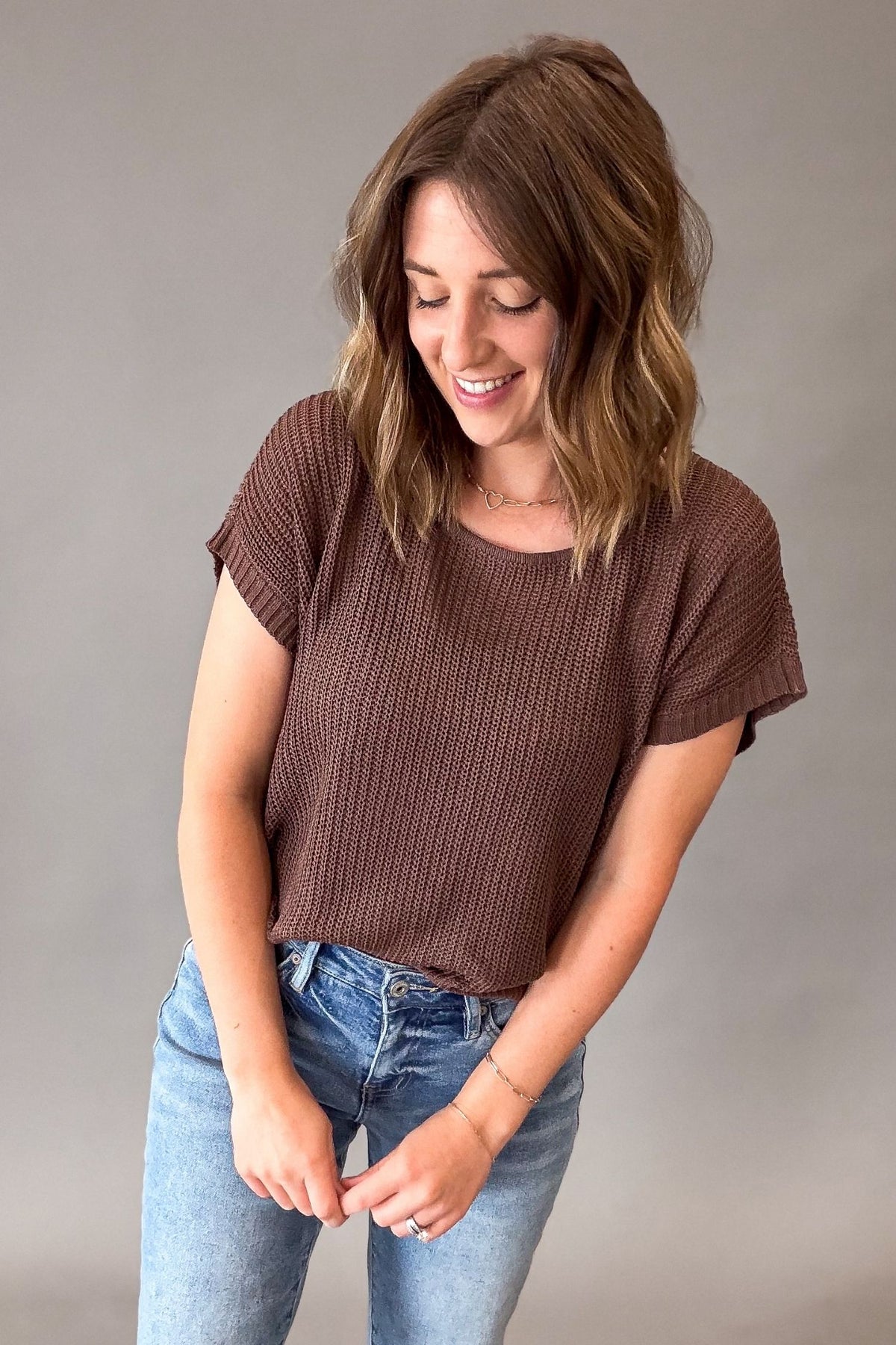 A woman wearing a brown short-sleeve textured knit top and light blue jeans, standing against a plain gray studio background.