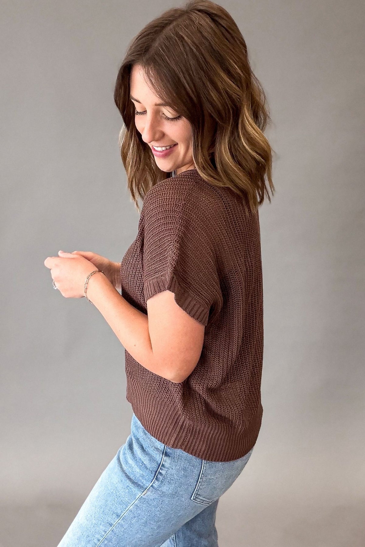 A woman wearing a brown short-sleeve knit top and light blue jeans, photographed against a plain gray studio background.
