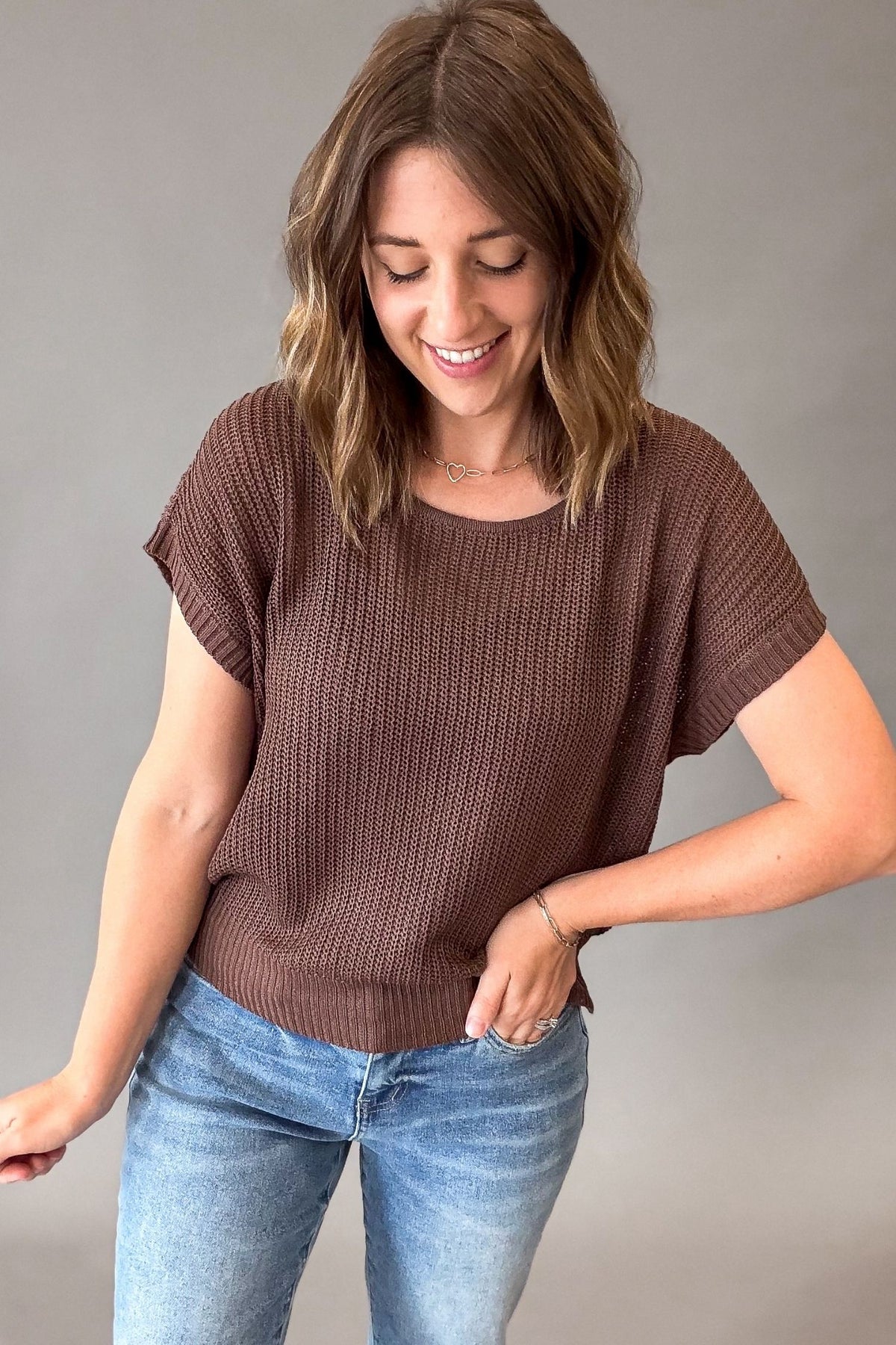 A woman wearing a brown short-sleeve ribbed knit top and blue jeans, standing against a plain gray studio background.