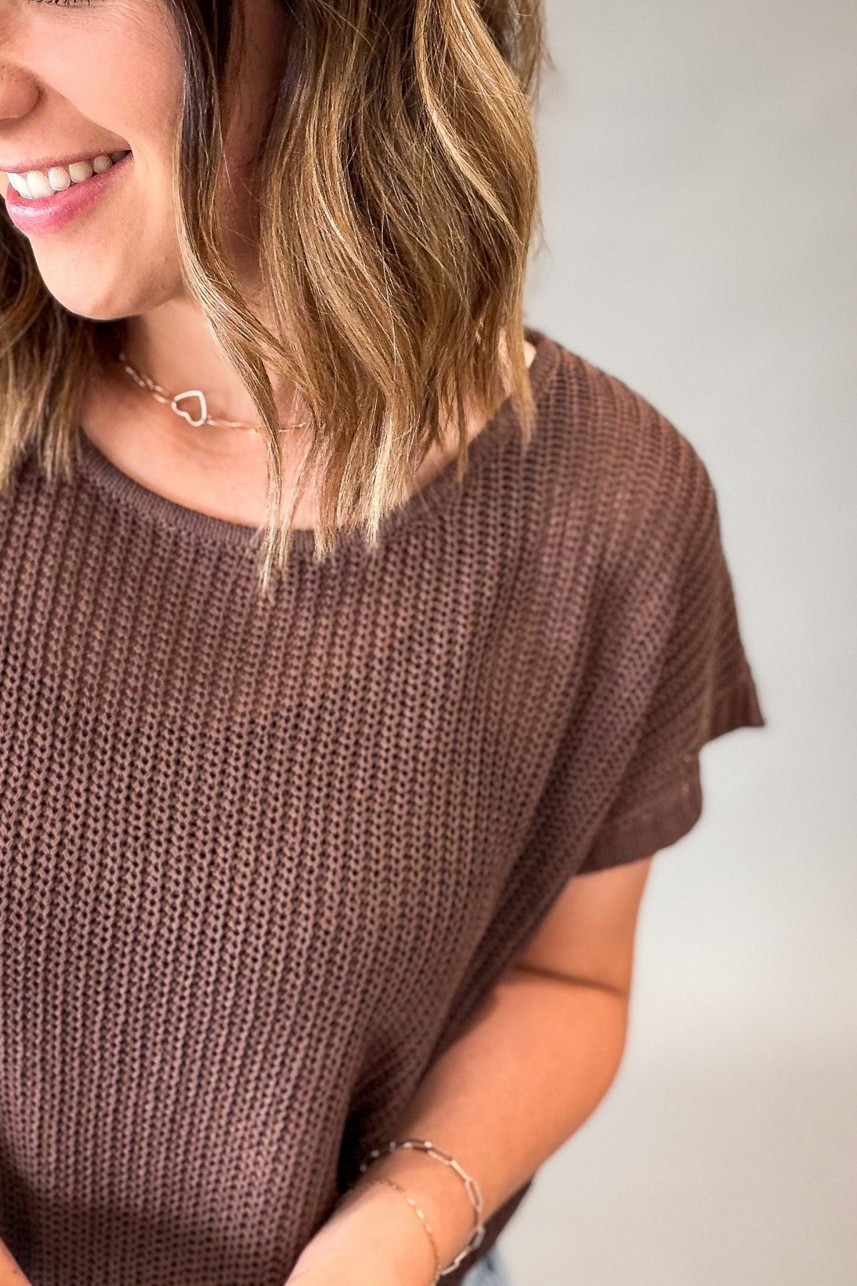 A close-up of a woman wearing a brown short-sleeve knit top, showing her smiling and hair against a plain light gray background.