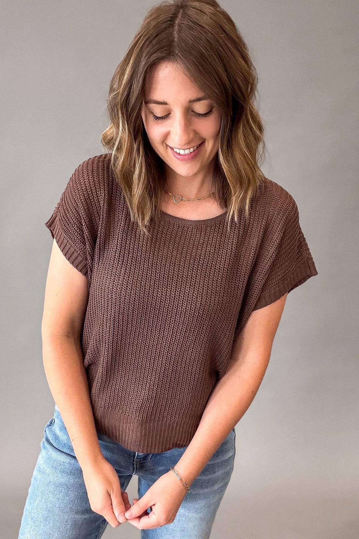 A woman wearing a brown short-sleeve knit top and blue jeans, standing against a plain gray studio background.