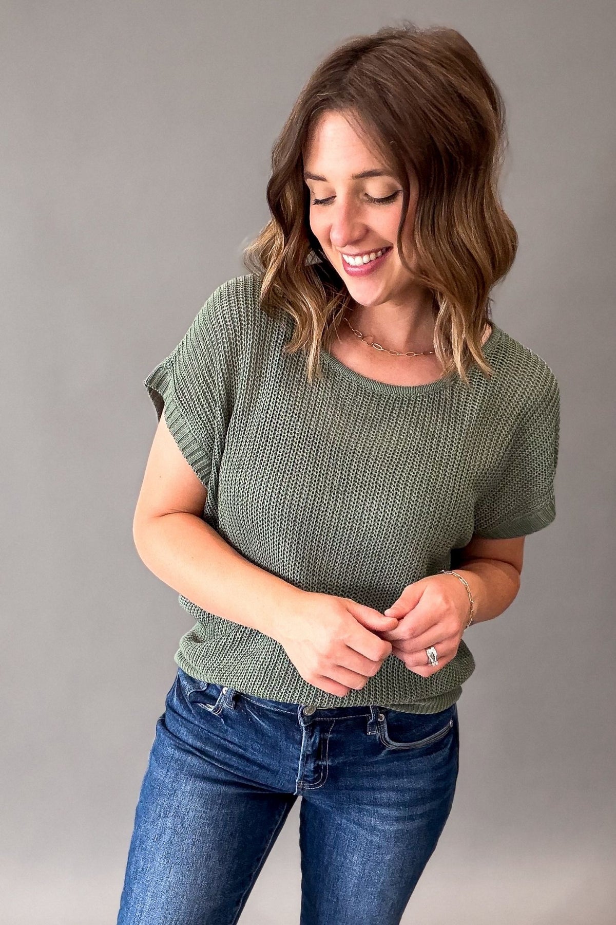 A woman wearing a textured emerald green short-sleeve knit top and blue jeans, standing against a solid gray studio background.