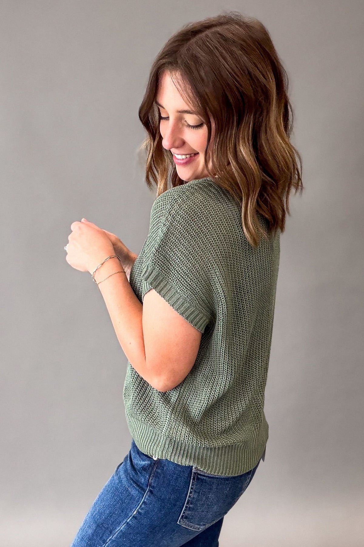 A woman with shoulder-length brown hair is shown from the back wearing a textured emerald green short-sleeve knit top and blue jeans against a plain gray studio background.