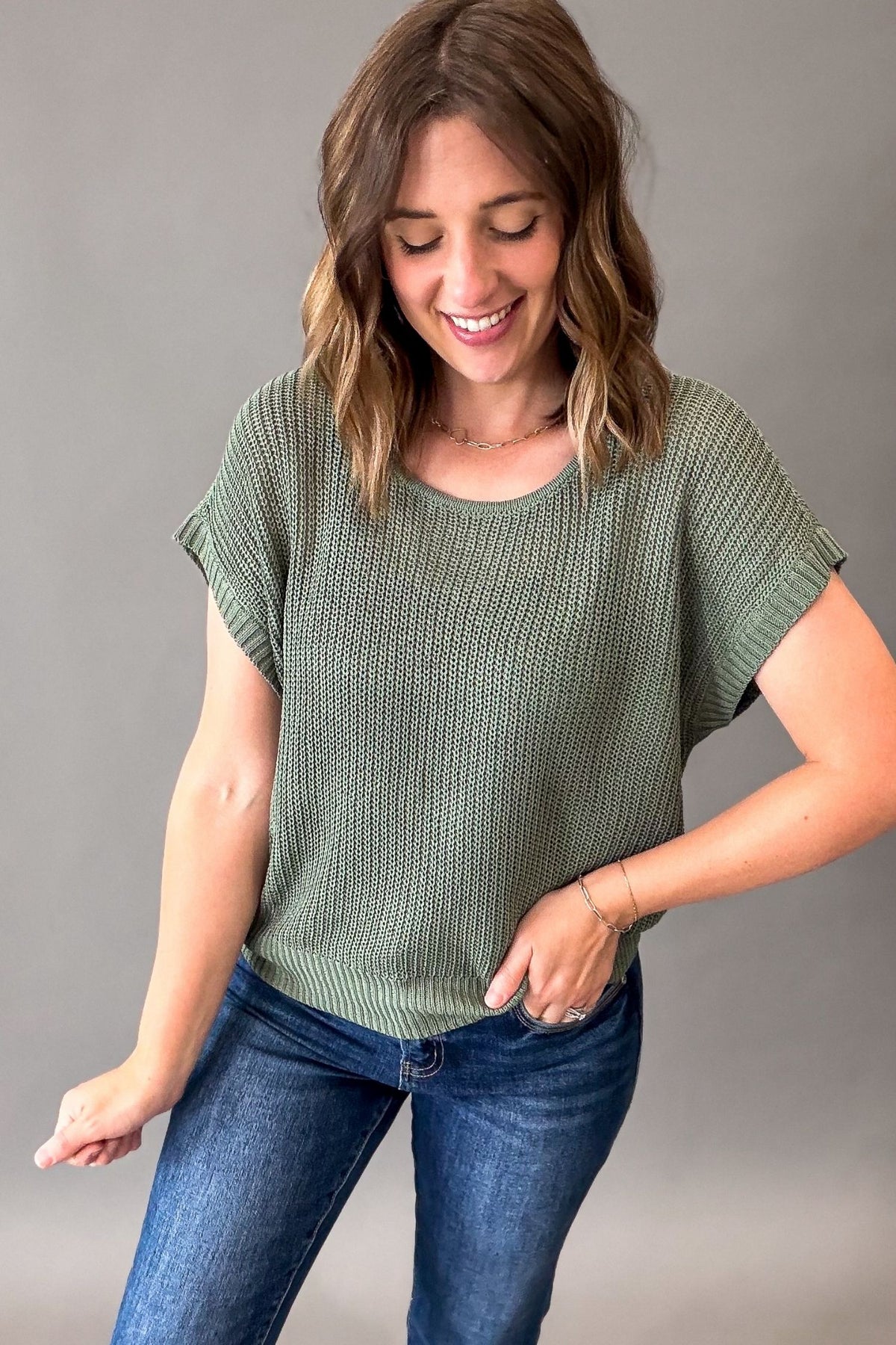 A woman wearing a textured emerald green short-sleeve top and blue jeans, posing against a neutral gray studio background.