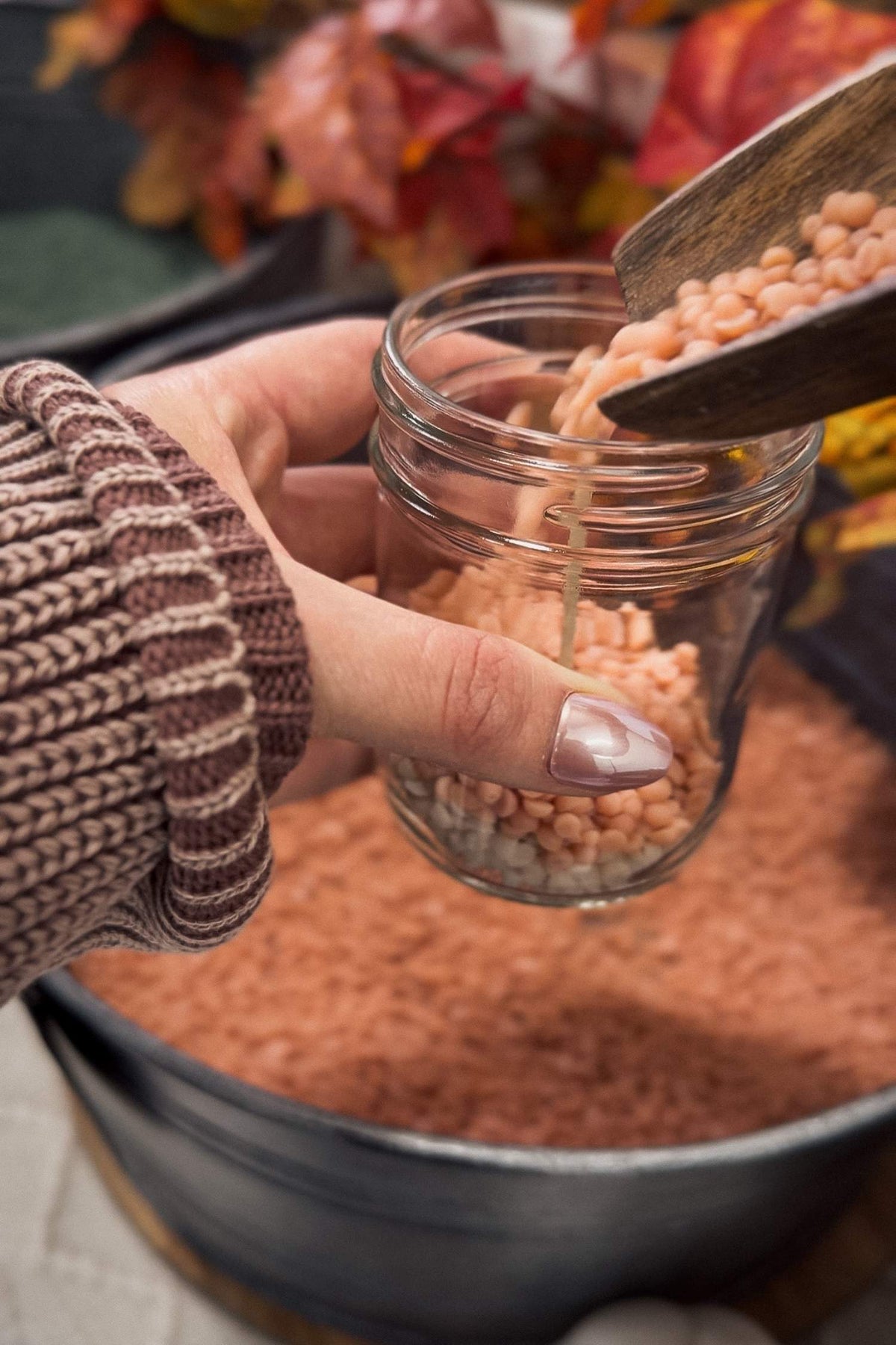 Hand holding a jar of orange beads with a wooden scoop, autumn leaves in the background
