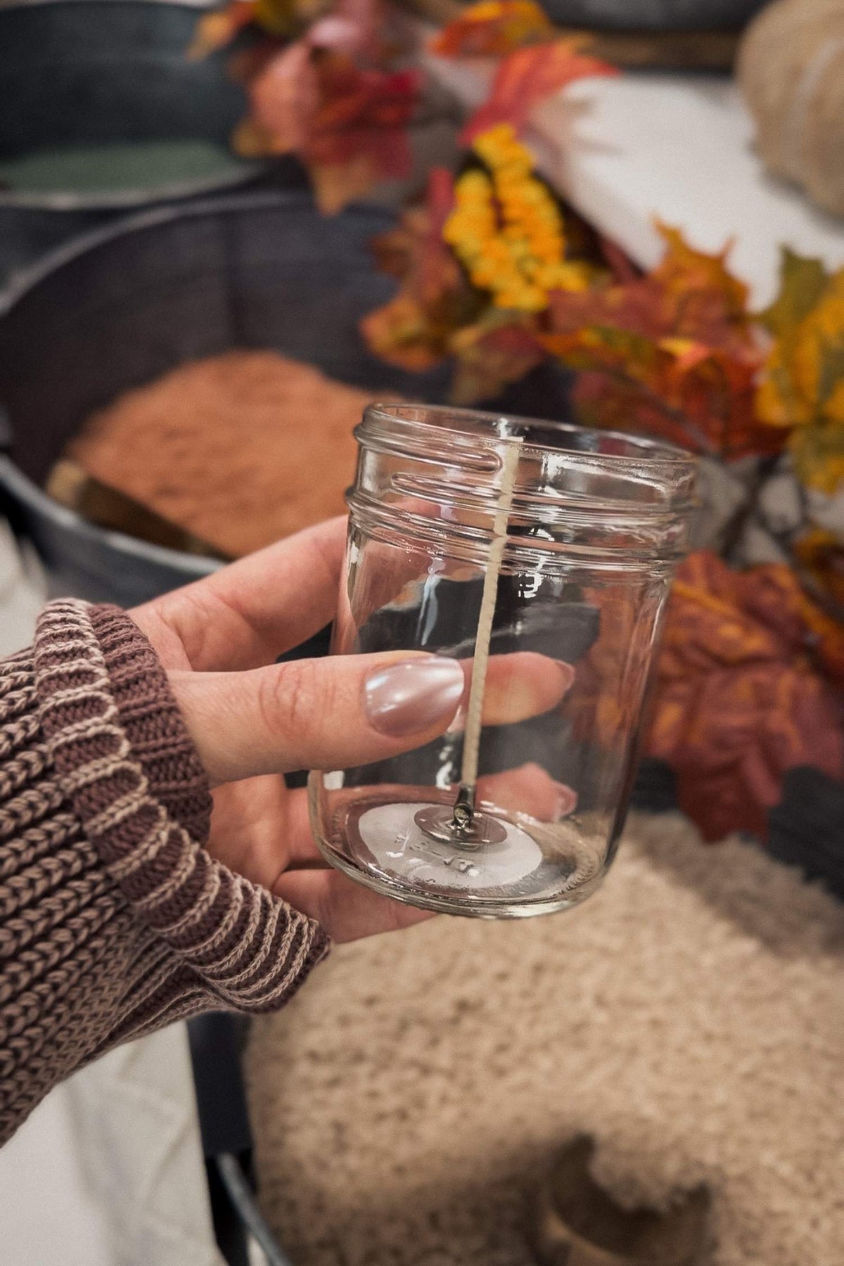 Hand holding a clear glass jar with a candle inside, surrounded by autumn leaves and decorations.