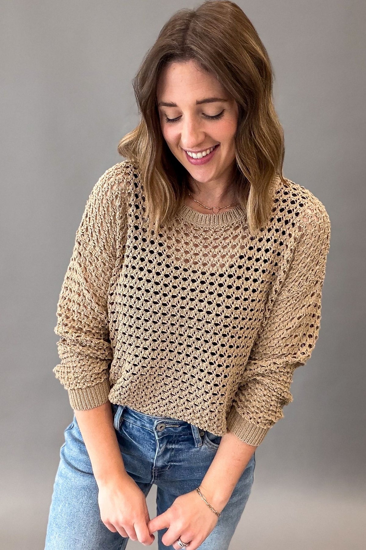 A woman wearing a latte colored open-knit crewneck sweater and blue jeans, smiling and looking down against a solid gray studio background.