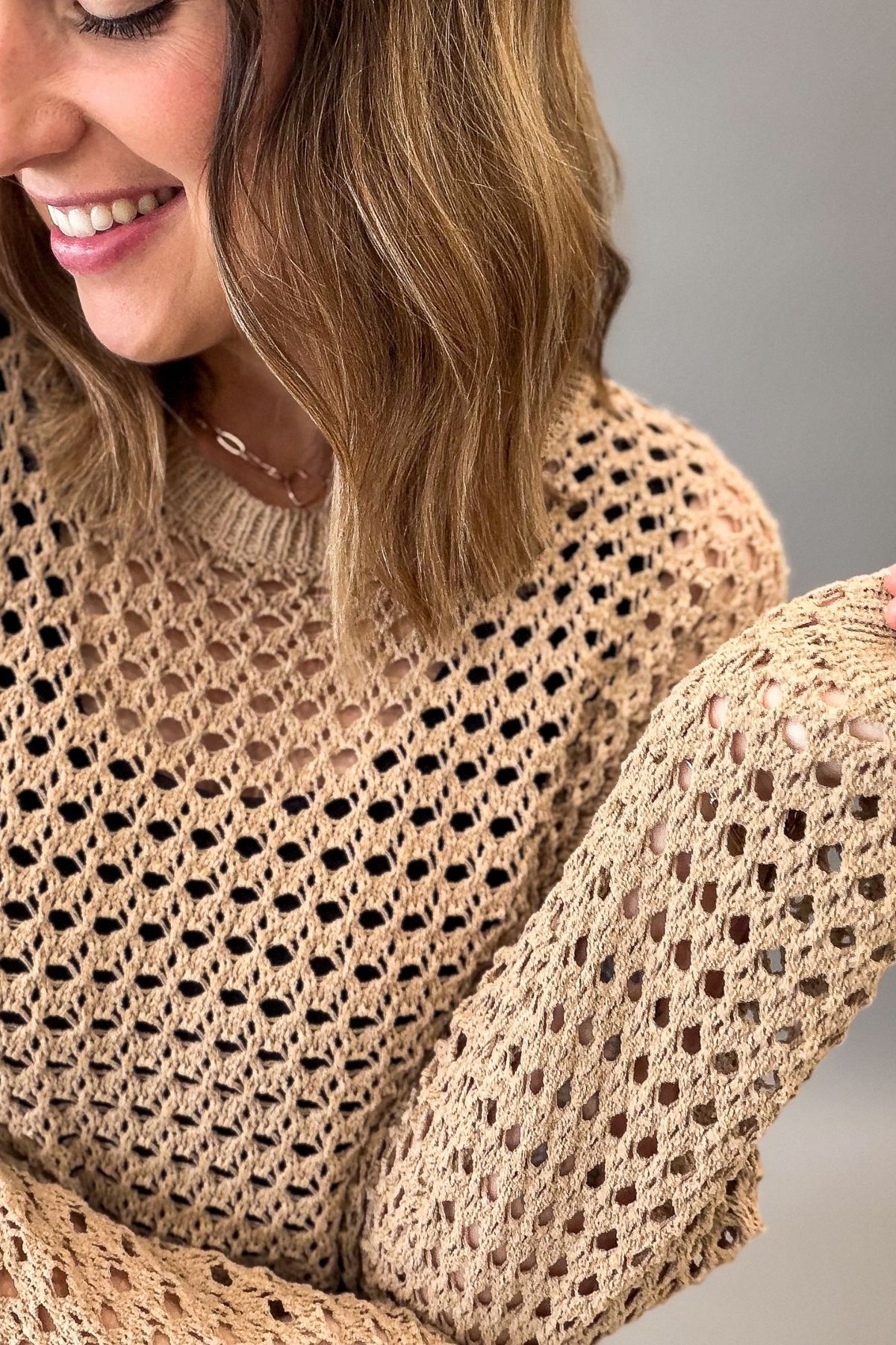 A close-up of a woman wearing a latte colored open-knit sweater with a crew neck and long sleeves, shown against a neutral gray background.