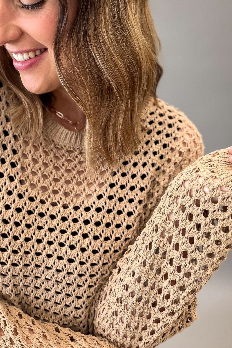 A close-up of a woman wearing a latte colored open-knit sweater with a crew neck and long sleeves, shown against a neutral gray background.