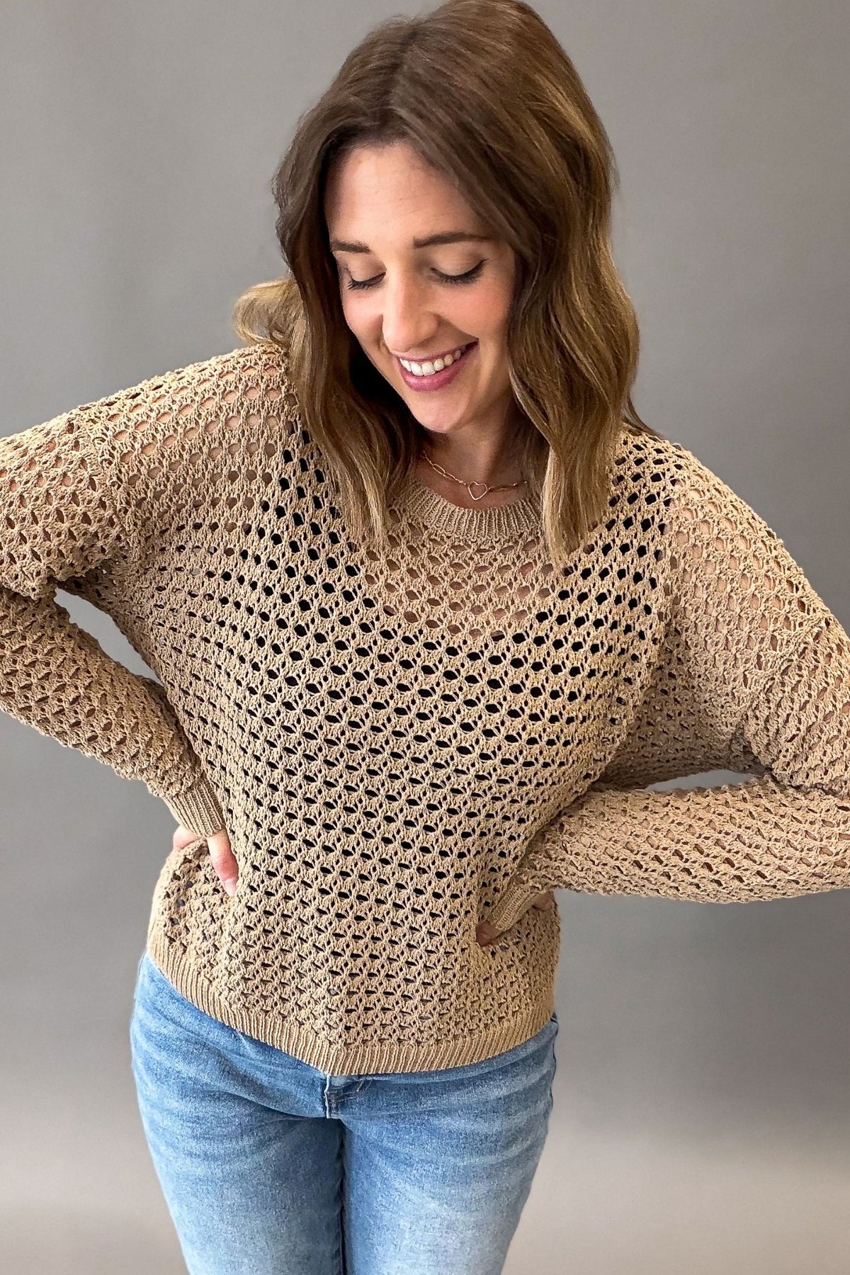 A woman wearing a latte colored open-knit crewneck sweater and blue jeans, posing with her hands on her hips against a solid gray studio background.