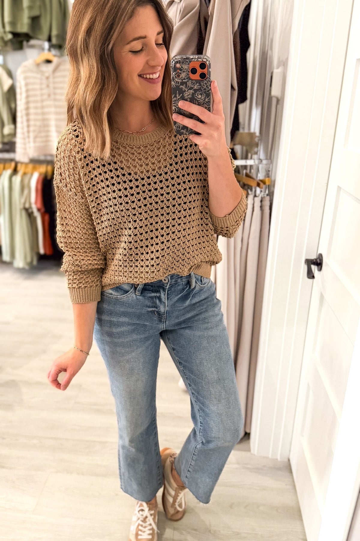 A woman takes a mirror selfie in a clothing boutique, wearing a latte colored open-knit crewneck sweater, light-wash blue jeans, and beige sneakers.