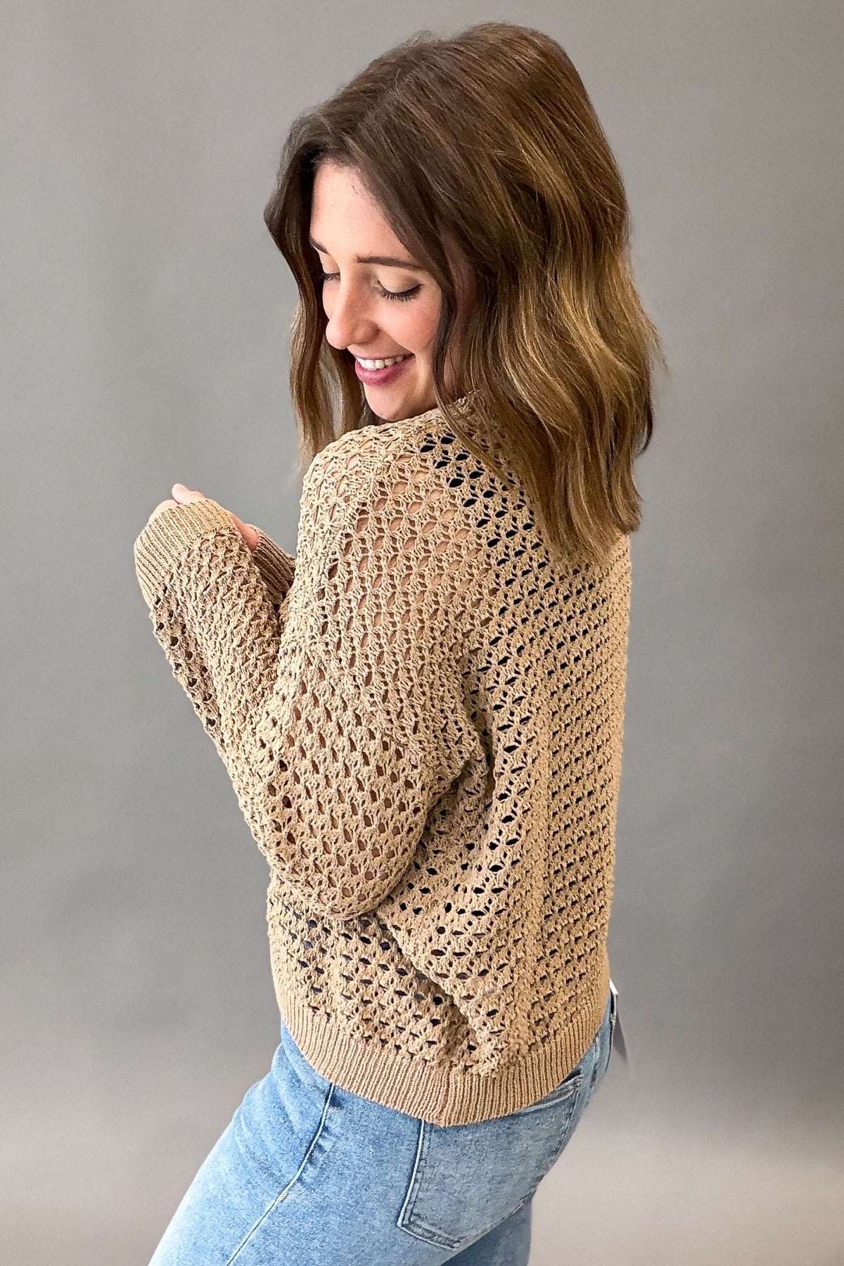 A woman wearing a late colored chunky open-knit sweater and light blue jeans, shown from a side-back angle against a plain gray studio background.