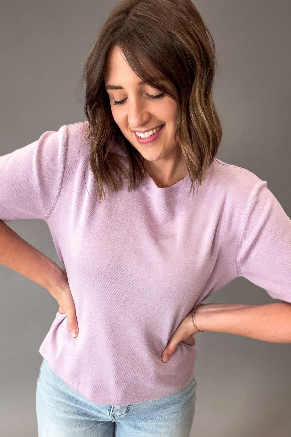 Woman wearing a lavender light weight sweater short sleeve and light blue jeans against a gray background